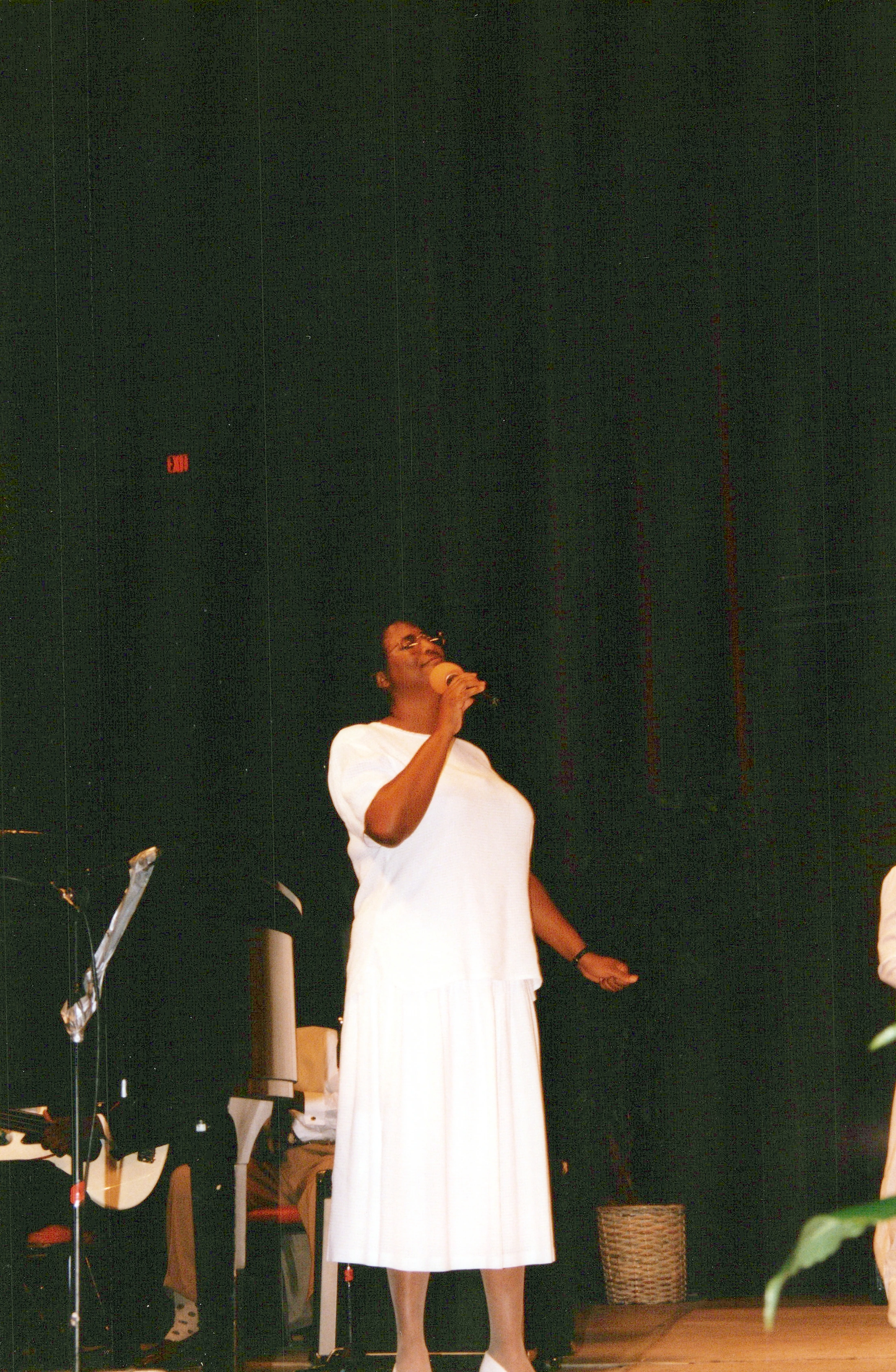 A Black woman in a white outfit stands on a stage, singing passionately into a microphone against a dark curtain backdrop.