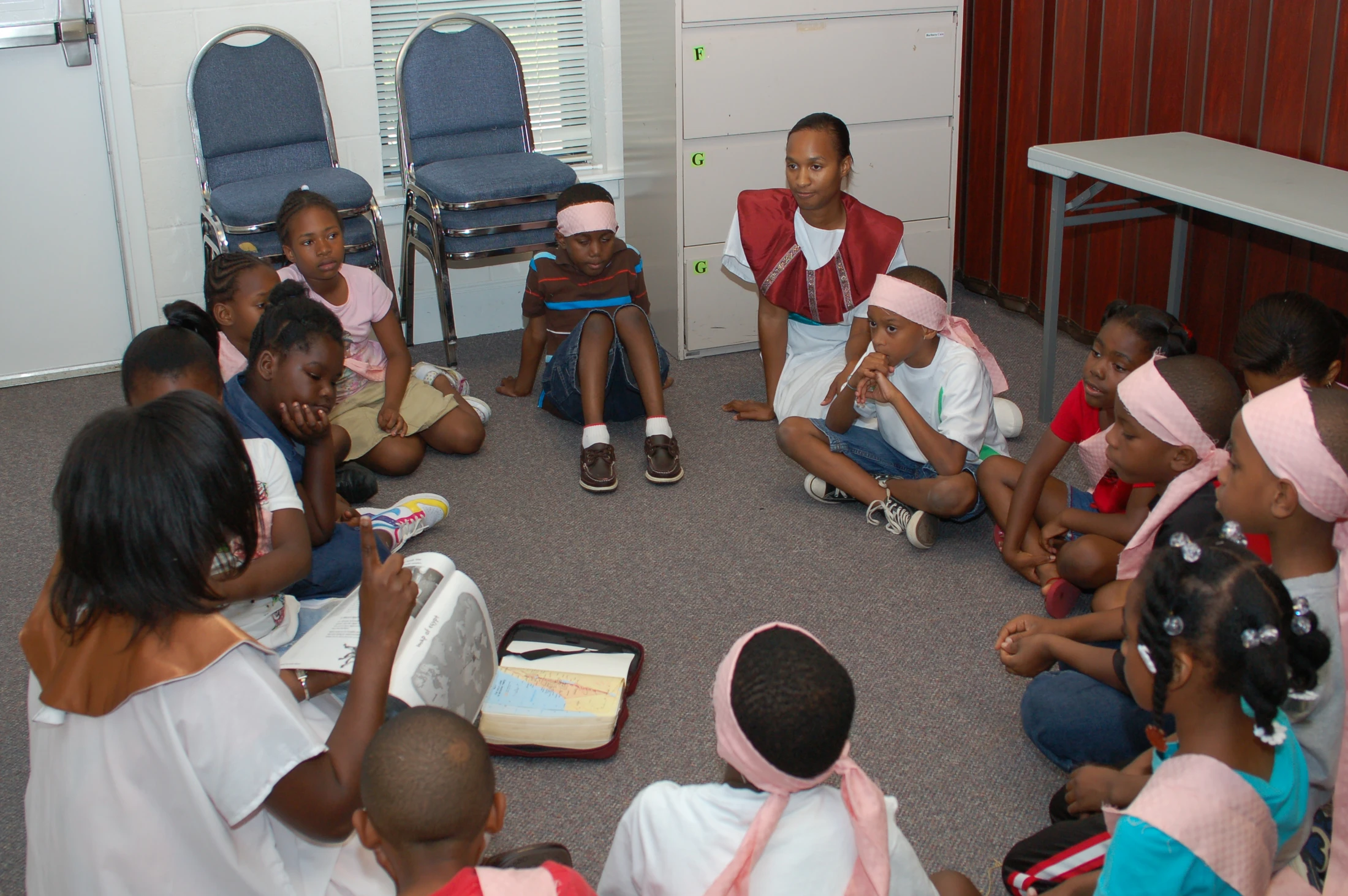 A group of Black children and two adults in simple costumes sit in a circle on the floor, listening attentively to a lesson from a book with maps.