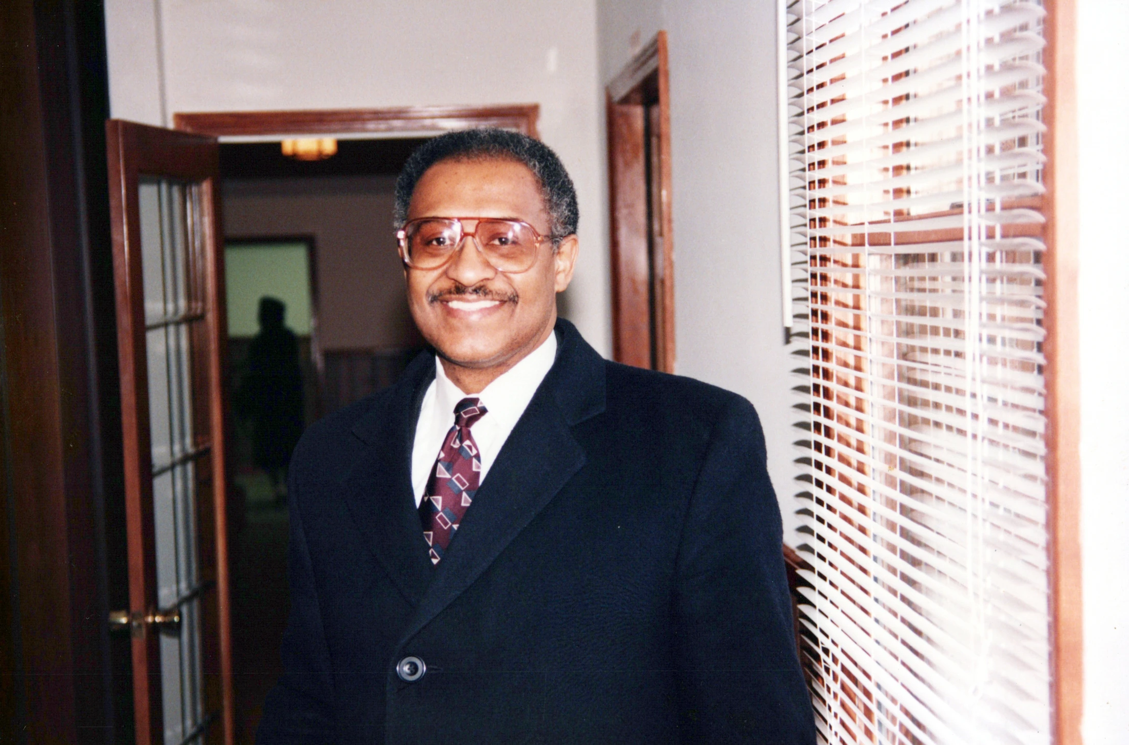 A medium shot of a smiling Black man with glasses and a mustache, wearing a dark overcoat, white shirt, and patterned tie, standing in an indoor hallway.