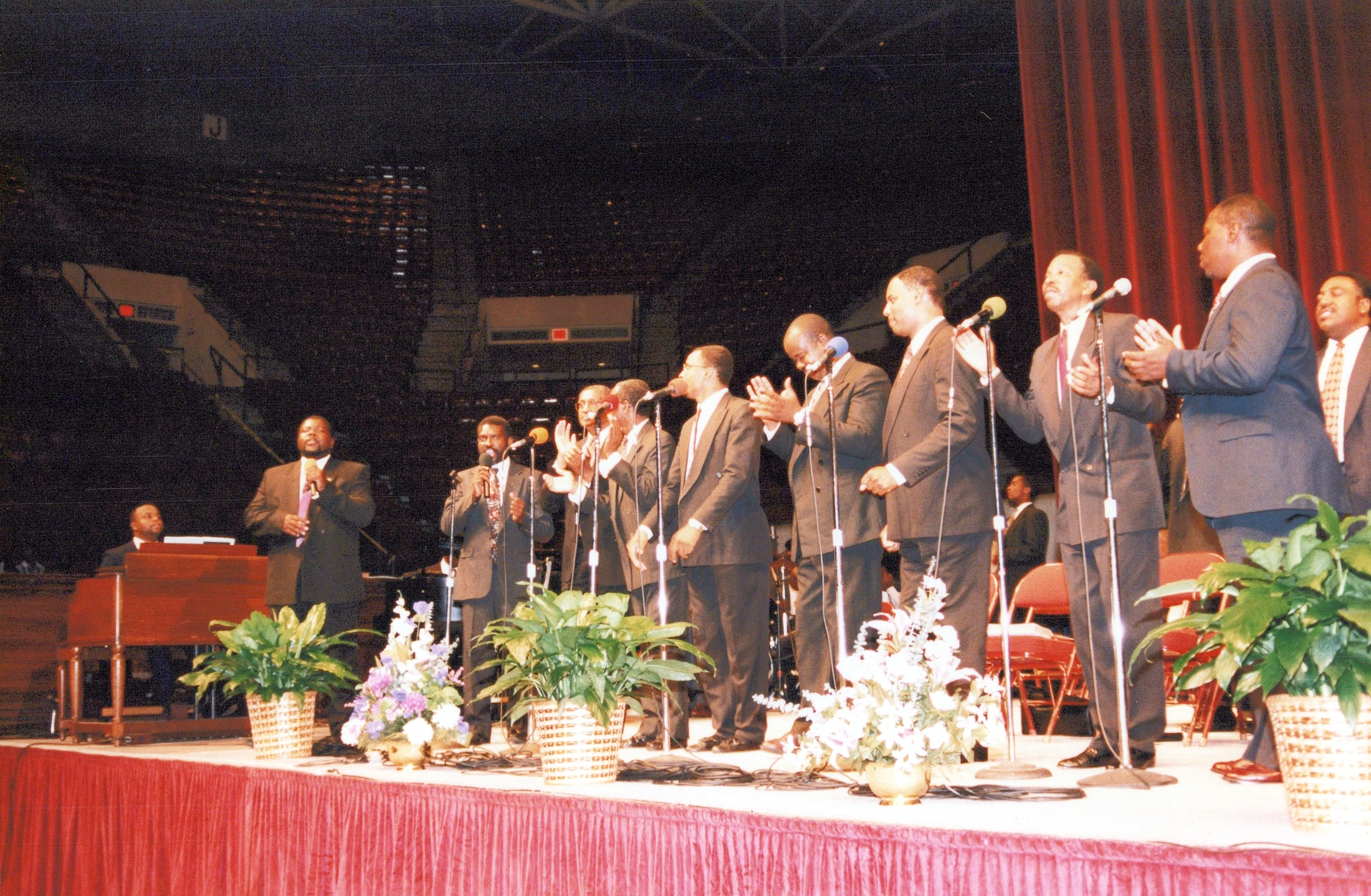 A line of African American men in dark suits sing and clap into microphones on a stage decorated with plants, with an organist to the left.