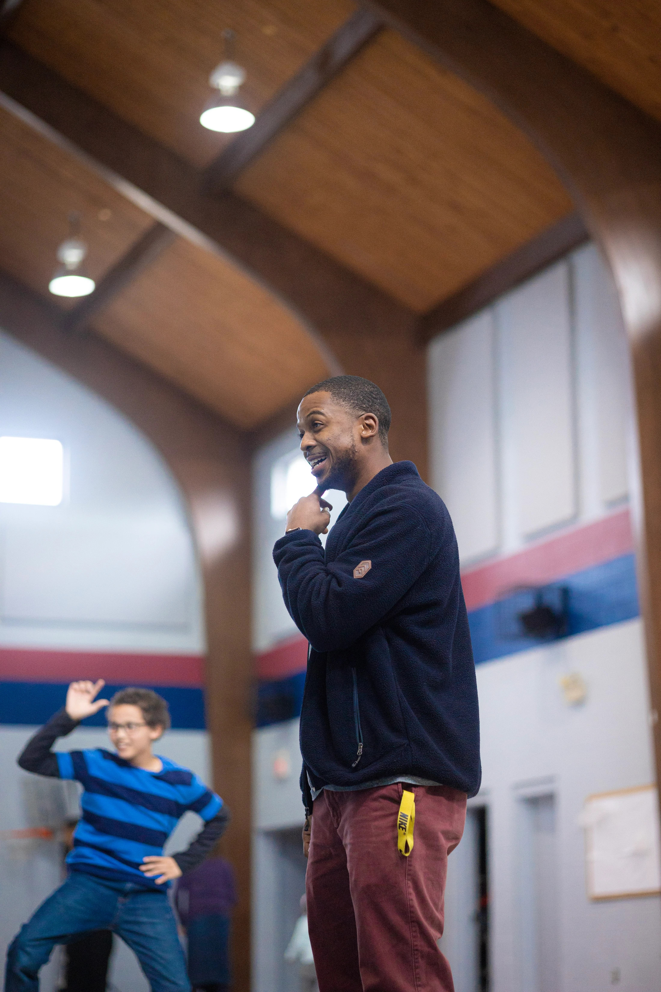 A Black man in a blue fleece jacket smiles while looking to his left. In the background, a young boy in a striped shirt is playing in a large hall.