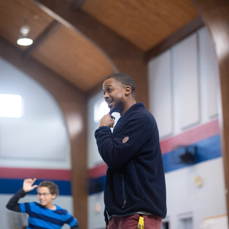 A Black man in a blue fleece jacket smiles while looking to his left. In the background, a young boy in a striped shirt is playing in a large hall.