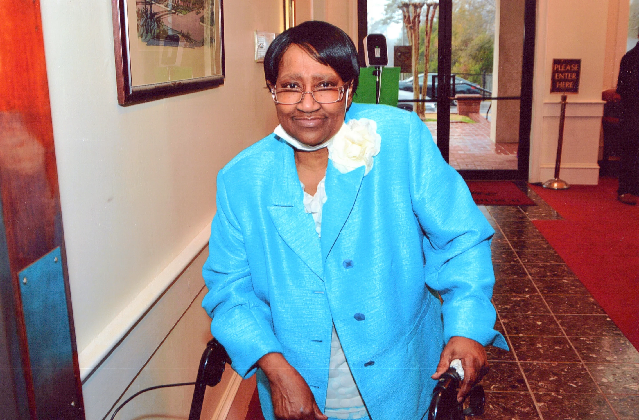 An elderly Black woman with glasses smiles warmly at the camera. She wears a bright blue jacket with a white flower corsage and is using a black walker.