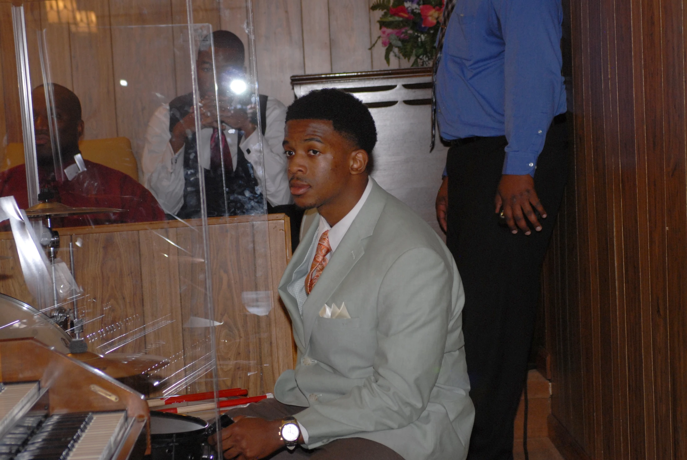 A young Black man in a light green suit and orange tie sits at a drum kit behind a clear shield, looking at the camera in a wood-paneled room.