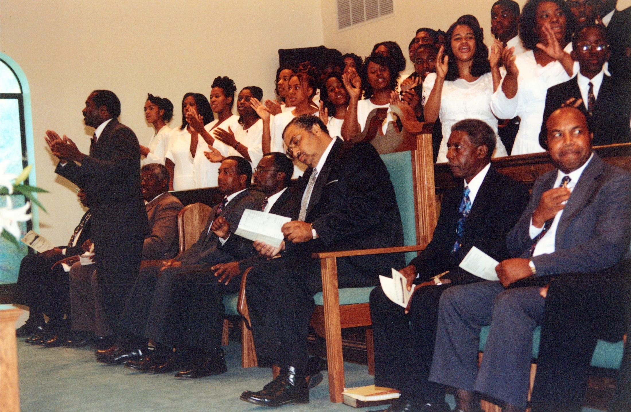A vintage photo of an African American church service, with men in suits seated in front pews and a choir standing, singing, and clapping in the background.