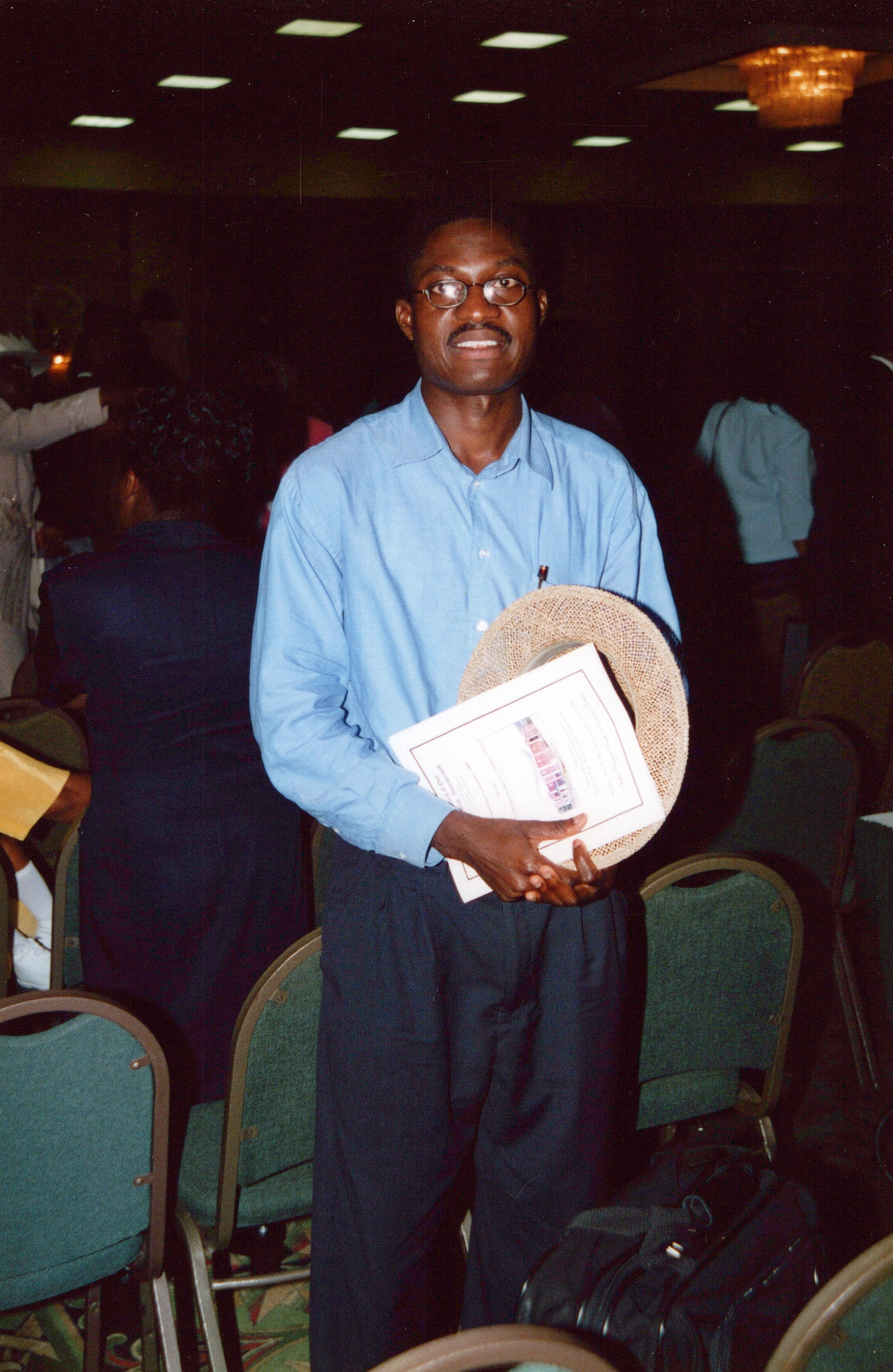 A man wearing glasses and a blue shirt smiles while standing in a conference room. He is holding a certificate and a straw hat.