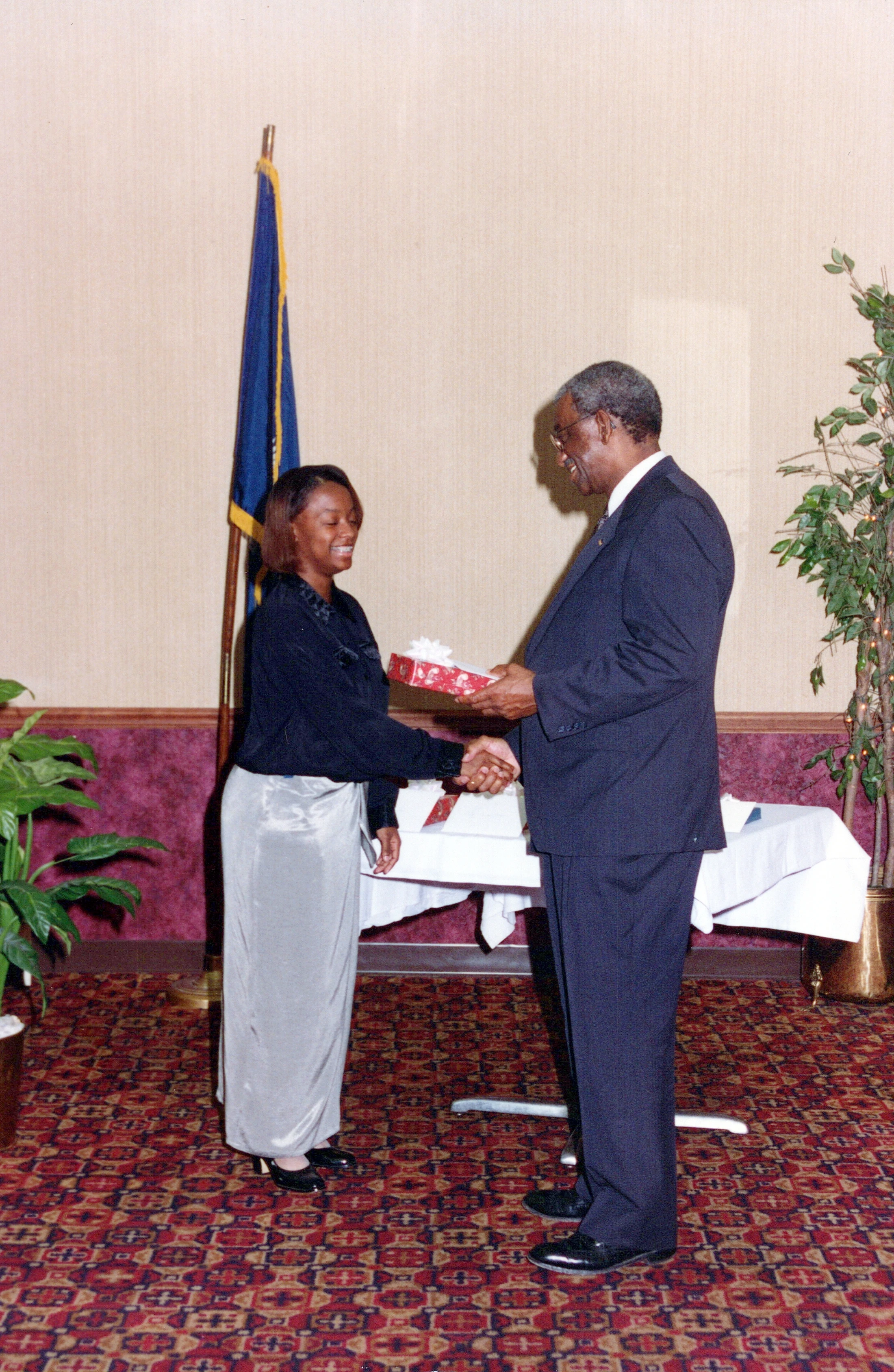 A smiling woman in a long skirt shakes hands with a man in a suit as he presents her with a gift box. They stand indoors in front of a flag.
