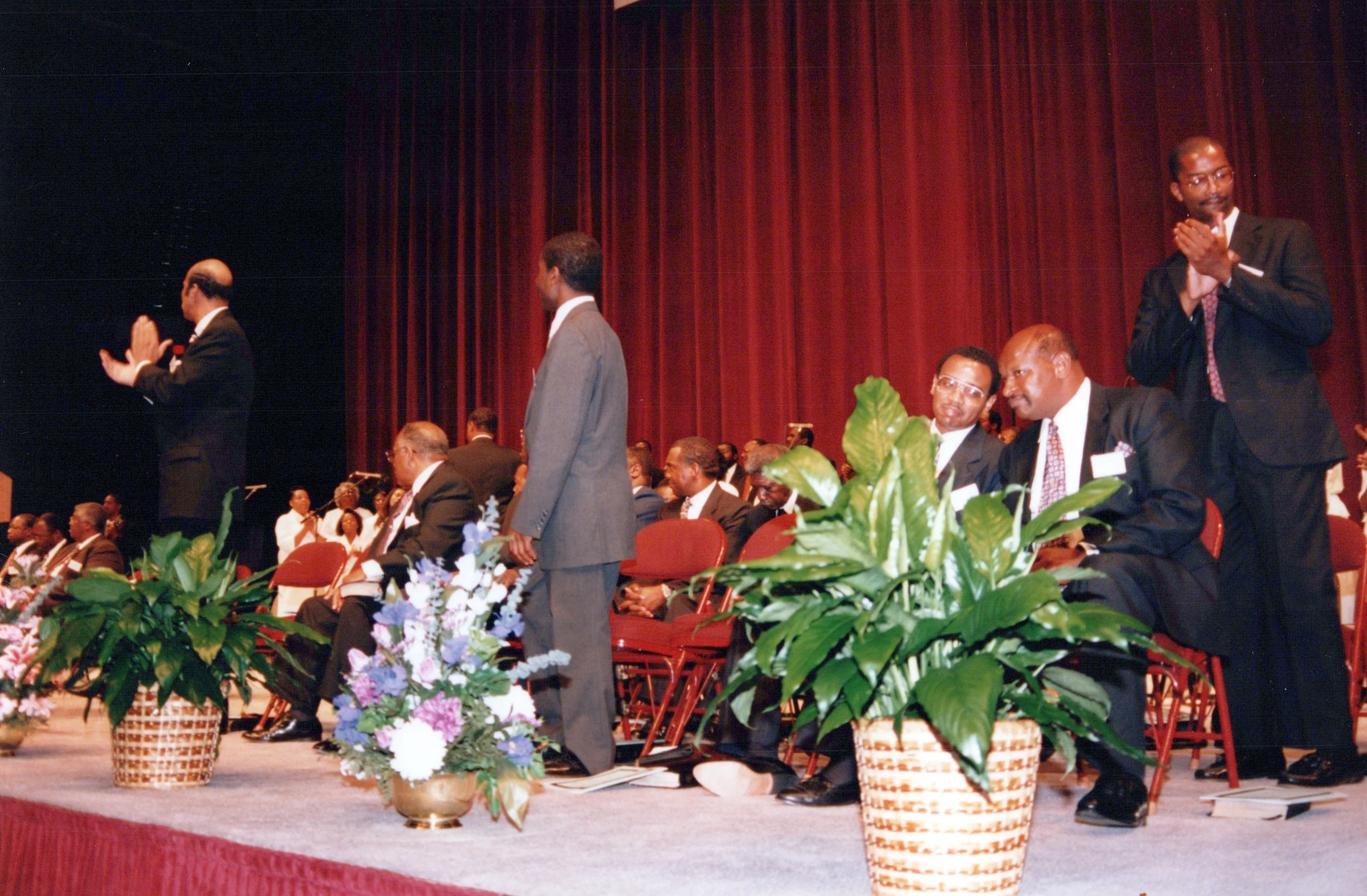 Several Black men in suits are on a stage with a red curtain. One man stands clapping, while others are seated or standing, watching a presentation or event.