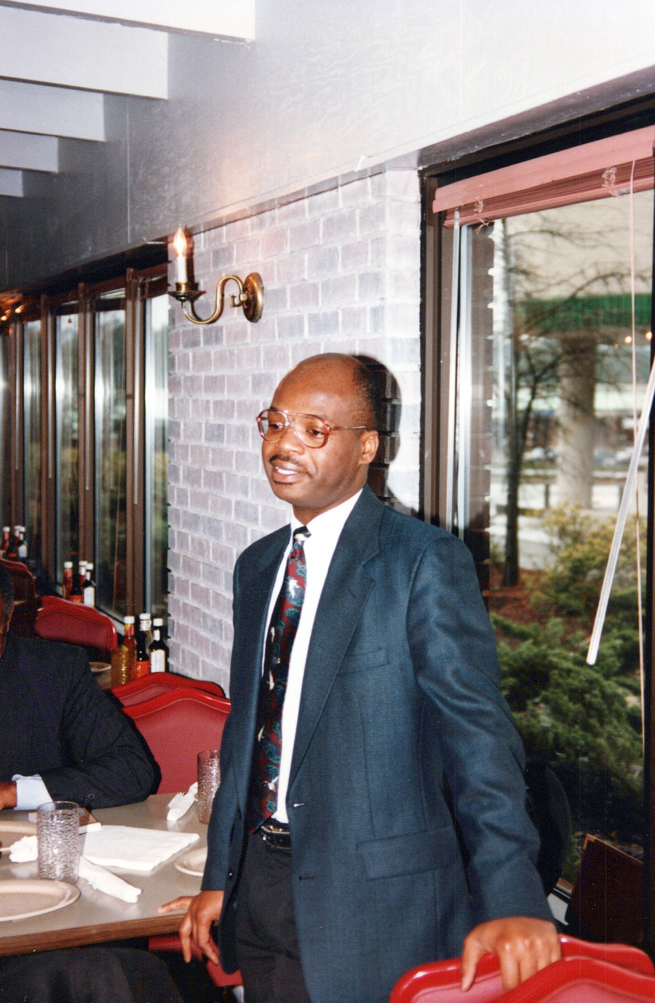 A Black man with glasses and a mustache, wearing a dark suit and patterned tie, stands smiling inside a restaurant by a table with red chairs.