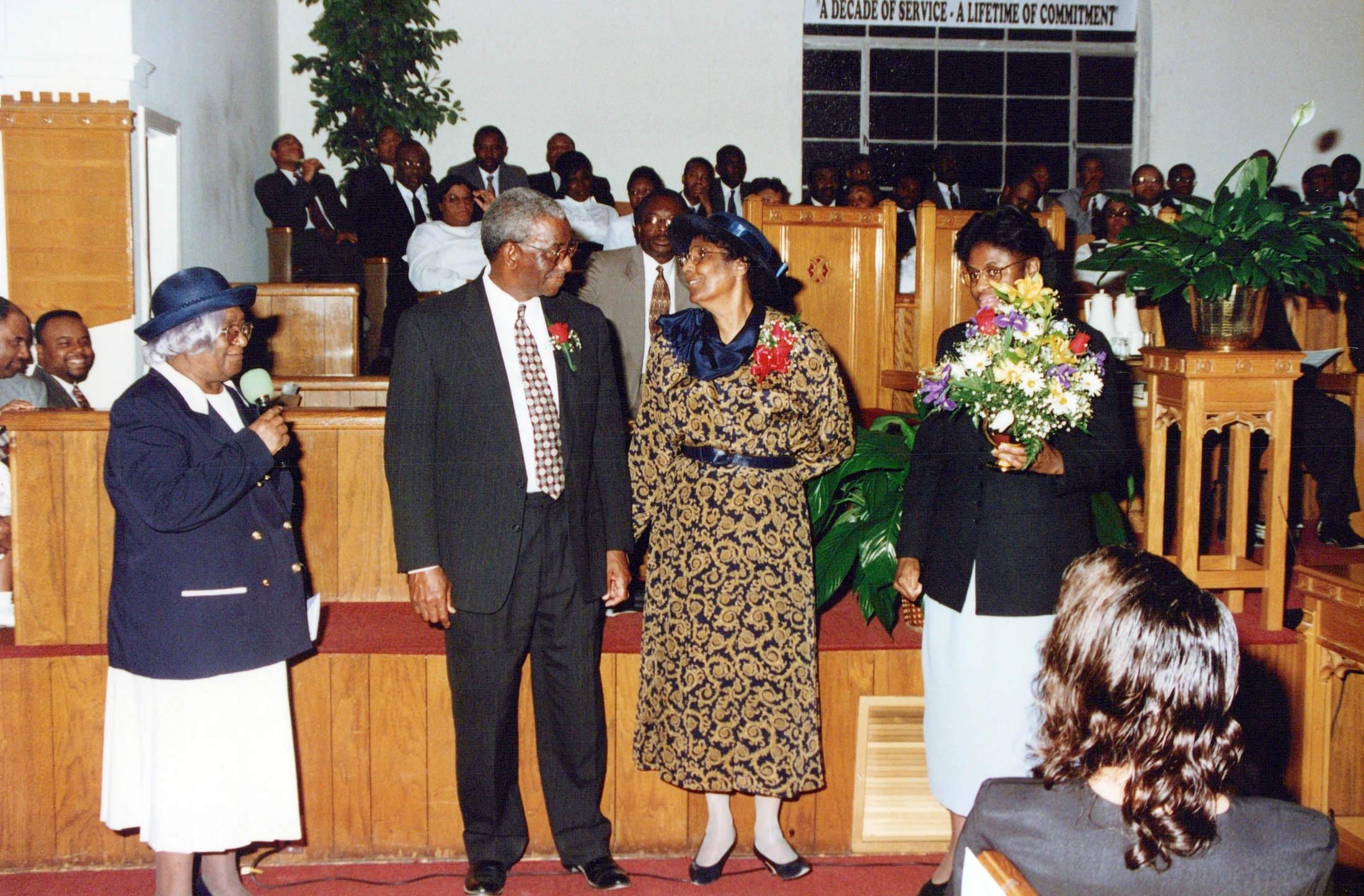 An older Black woman in a blue hat speaks at a church ceremony, standing with a man and two other women, one holding flowers, in front of a congregation.
