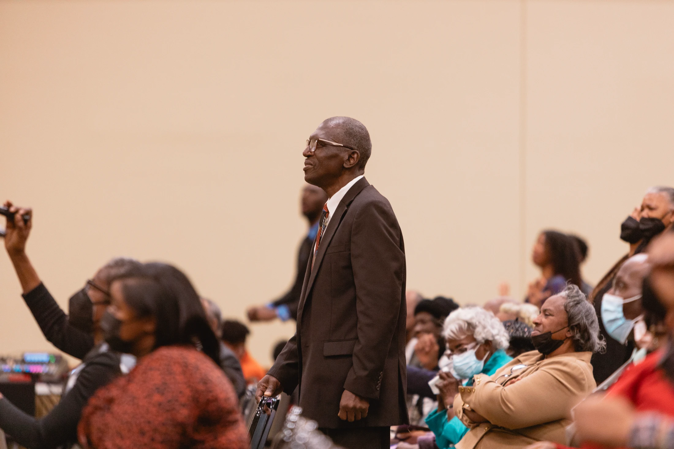 An elderly Black man in a dark suit and glasses stands in the middle of a seated congregation, looking upward with a thoughtful expression.
