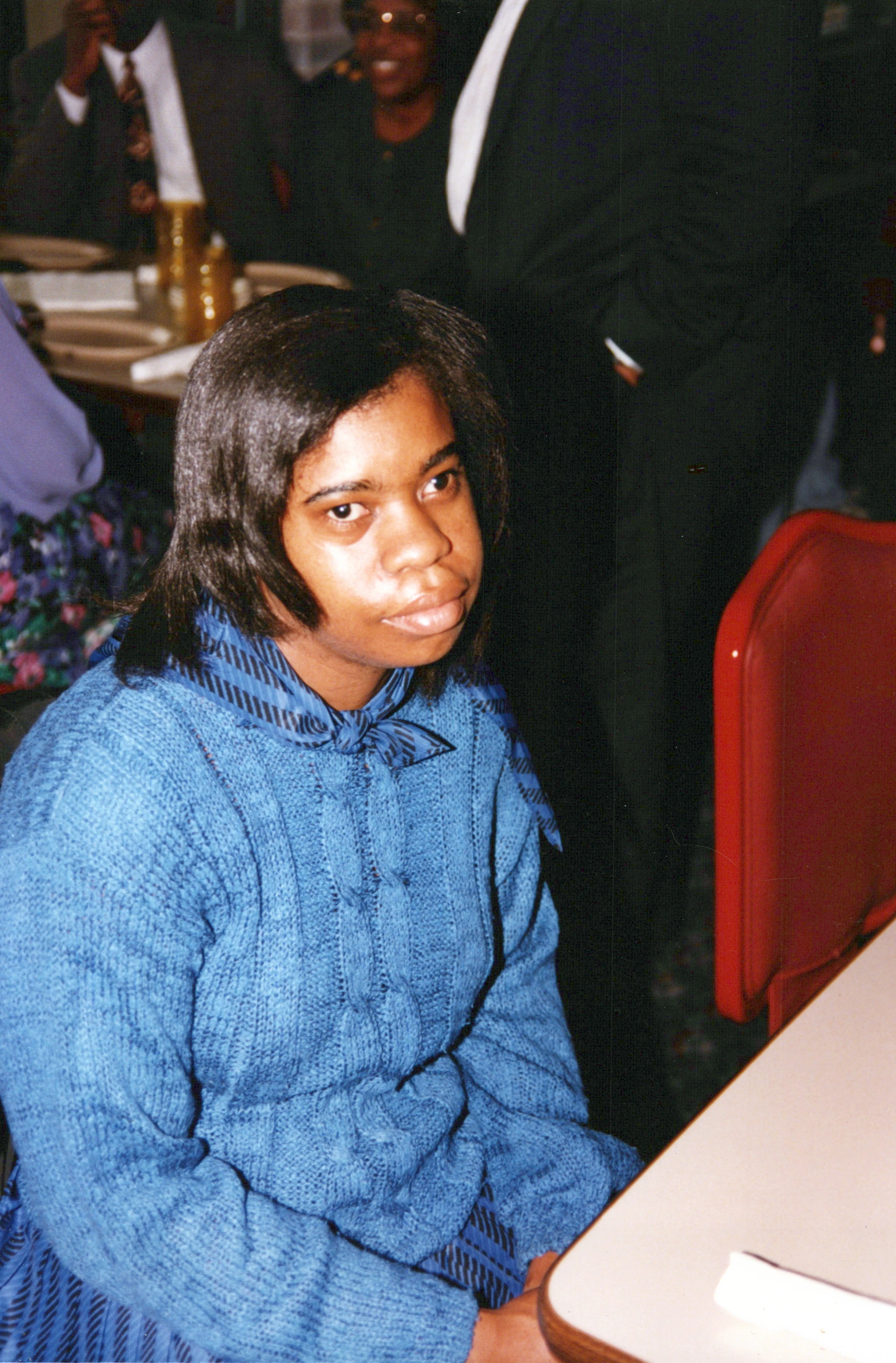 A vintage photograph of a Black woman wearing a blue cable-knit sweater and a patterned scarf, sitting at a table and looking to her right.