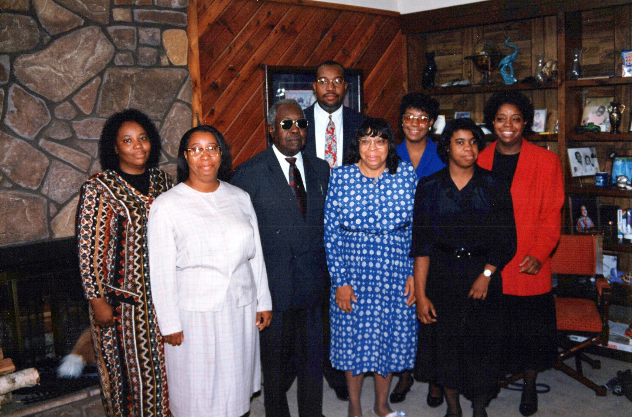 A vintage photograph of a large African American family, dressed up and smiling for a portrait in a living room with a stone fireplace and wood paneling.