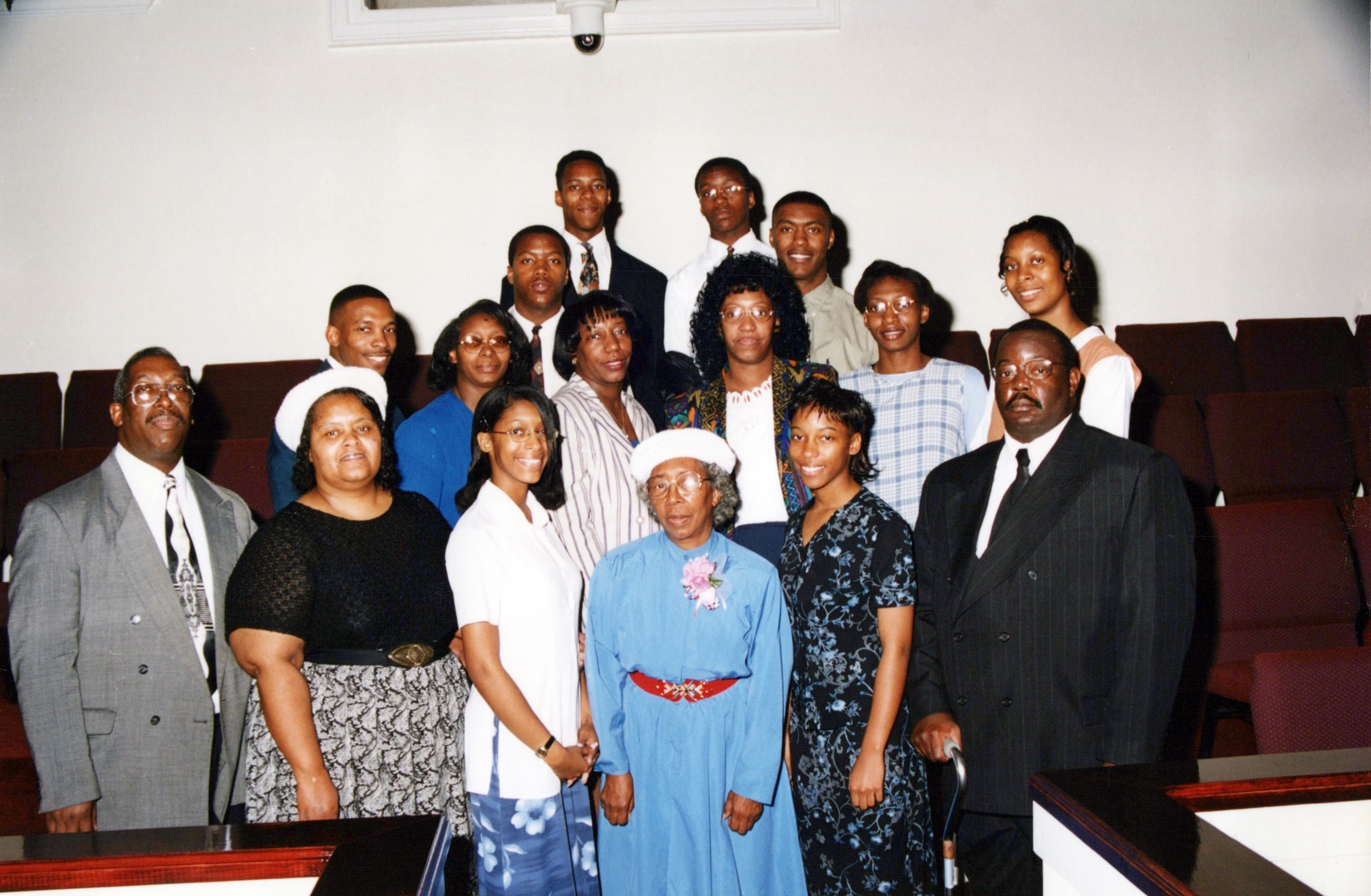 A large group of African American men, women, and teenagers pose for a formal photo inside a church, with an elderly woman in blue at the center.