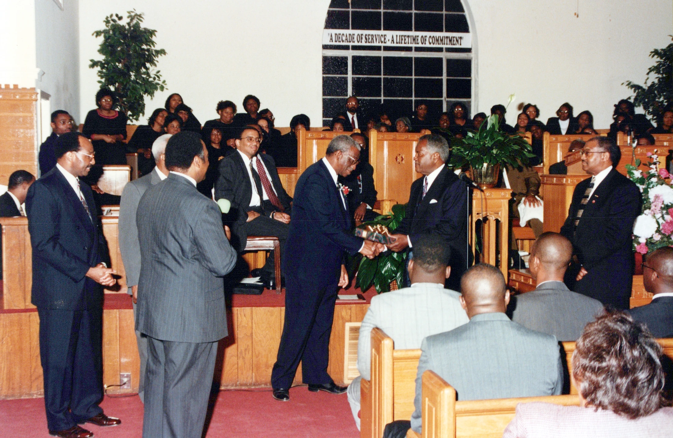 Men in suits stand at the front of a church during a service. One man presents a gift to another as the congregation watches from wooden pews.