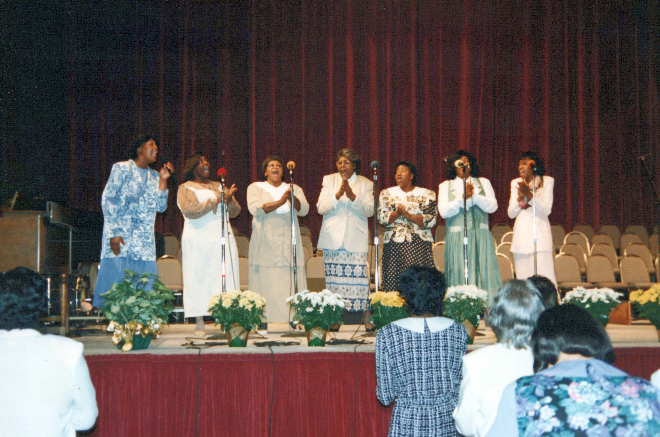 Seven African American women stand in a line on a stage, singing joyfully into microphones for an audience. They are in front of a red curtain.