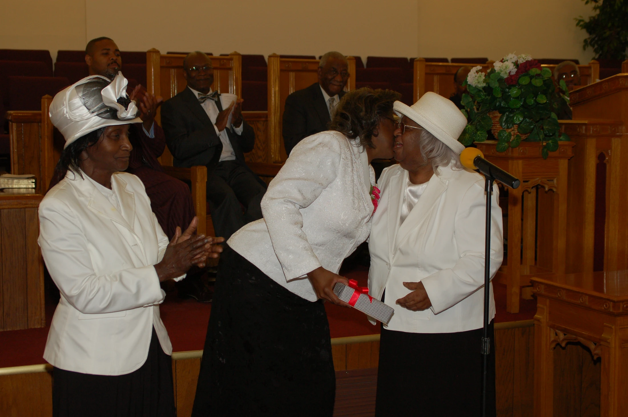 Two Black women in white suits and hats embrace in a church. One kisses the other's cheek while holding a gift, as other congregants clap in the pews.