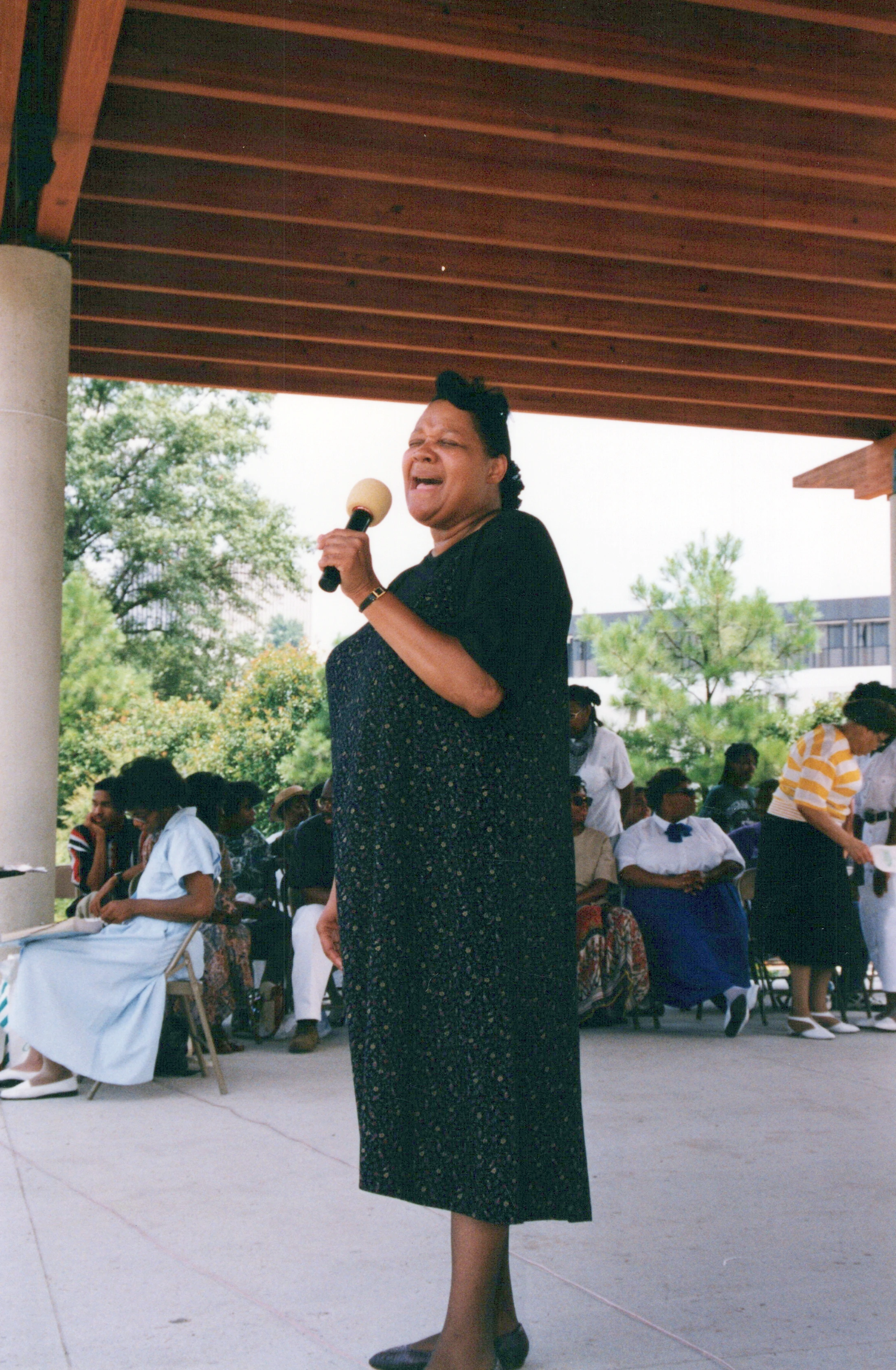 A Black woman in a long, dark patterned dress stands and sings into a microphone under a covered patio. An audience is seated in the background.