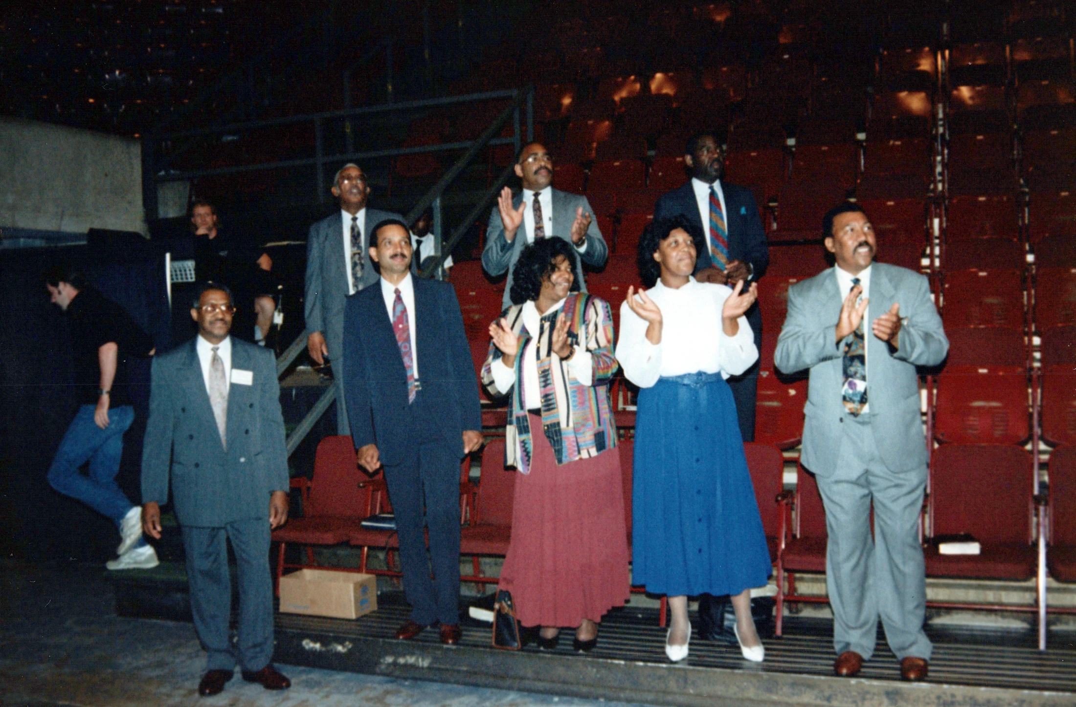 A vintage photograph shows a group of well-dressed Black men and women standing and applauding in a dimly lit auditorium with red seats.