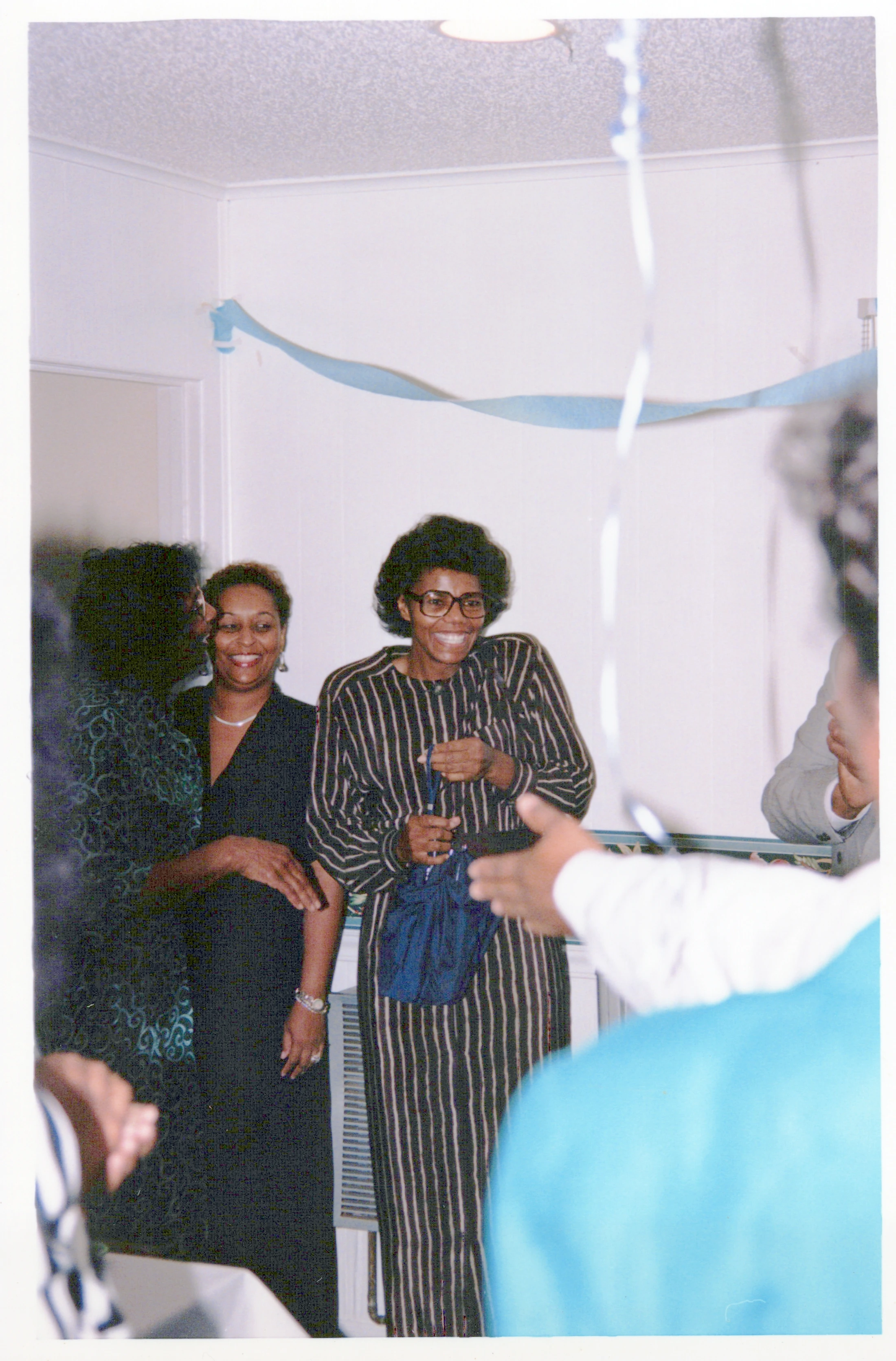 A vintage photograph shows three Black women smiling happily at an indoor party. The woman in the center wears glasses and a black and white striped dress.