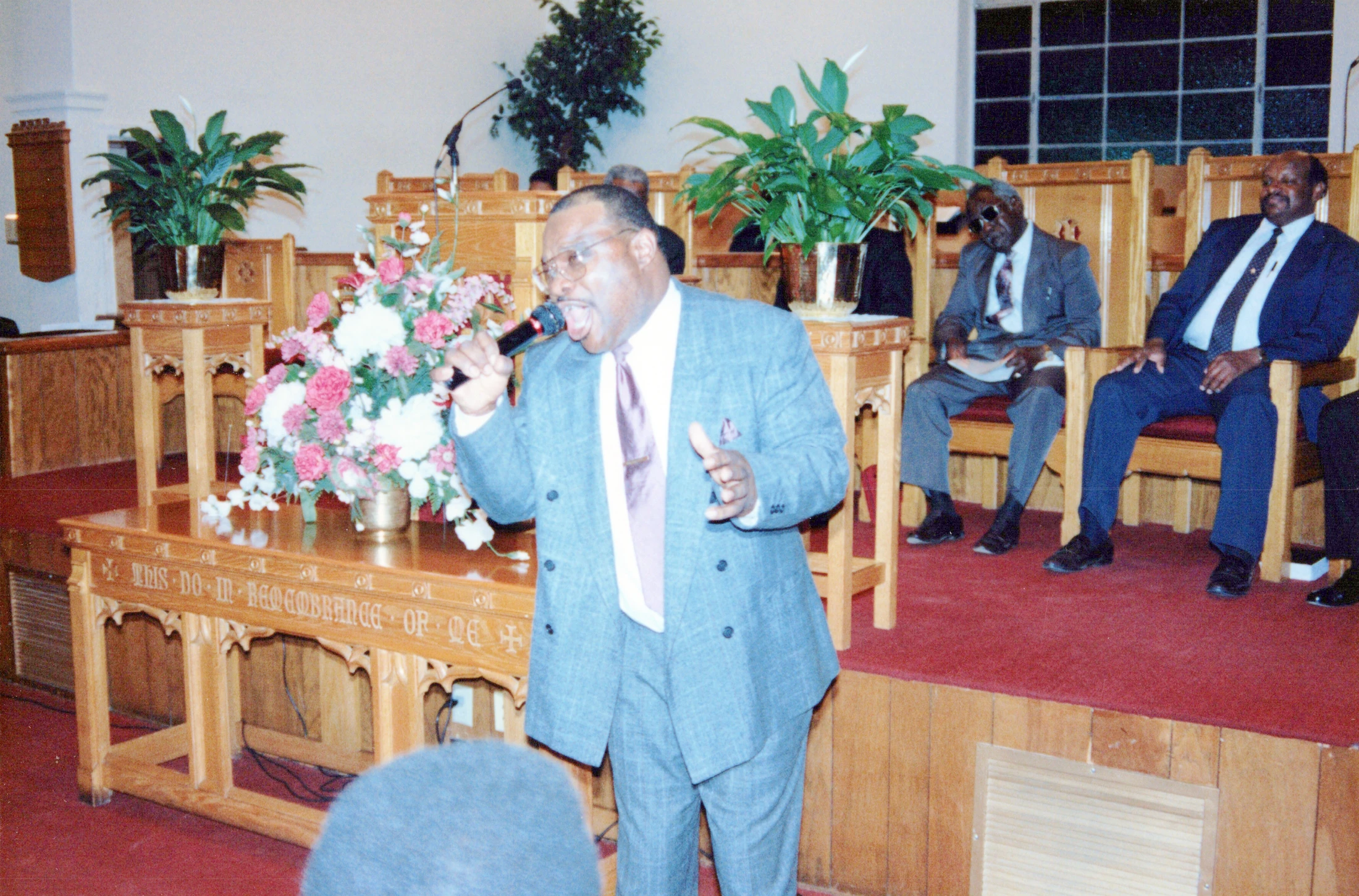 A Black man in a blue suit sings into a microphone during a church service. Two other men sit in suits behind him on a wooden altar with flowers.