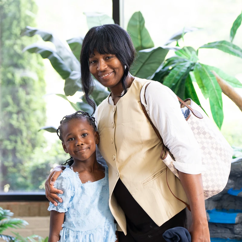 A smiling Black woman with her arm around a young Black girl in a blue dress. They are posing indoors in front of a large window with green plants.