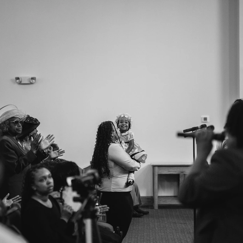 A black and white photo of a woman holding a smiling toddler in a bonnet. They are surrounded by a congregation of people who are clapping.