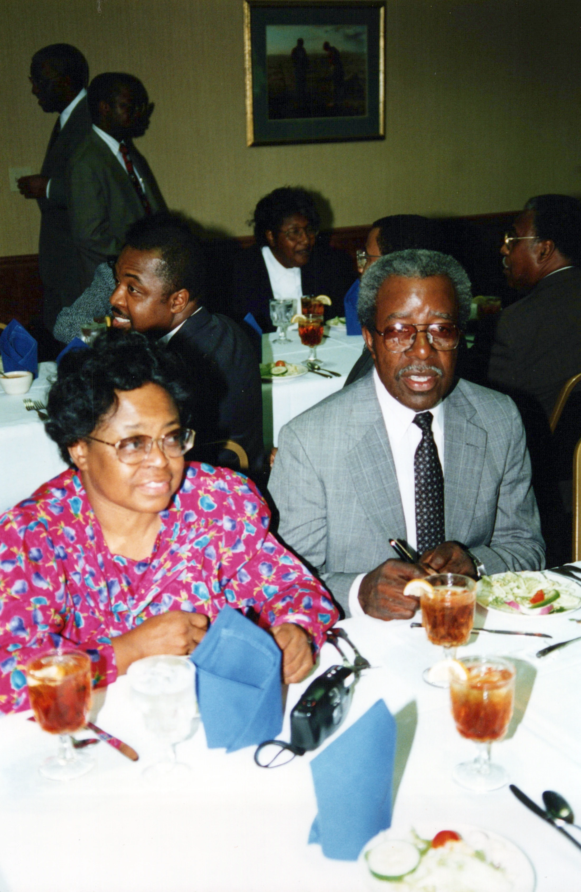 An older Black couple sits at a formal dinner table. The man wears a gray suit and the woman a colorful floral blouse. Other guests are in the background.