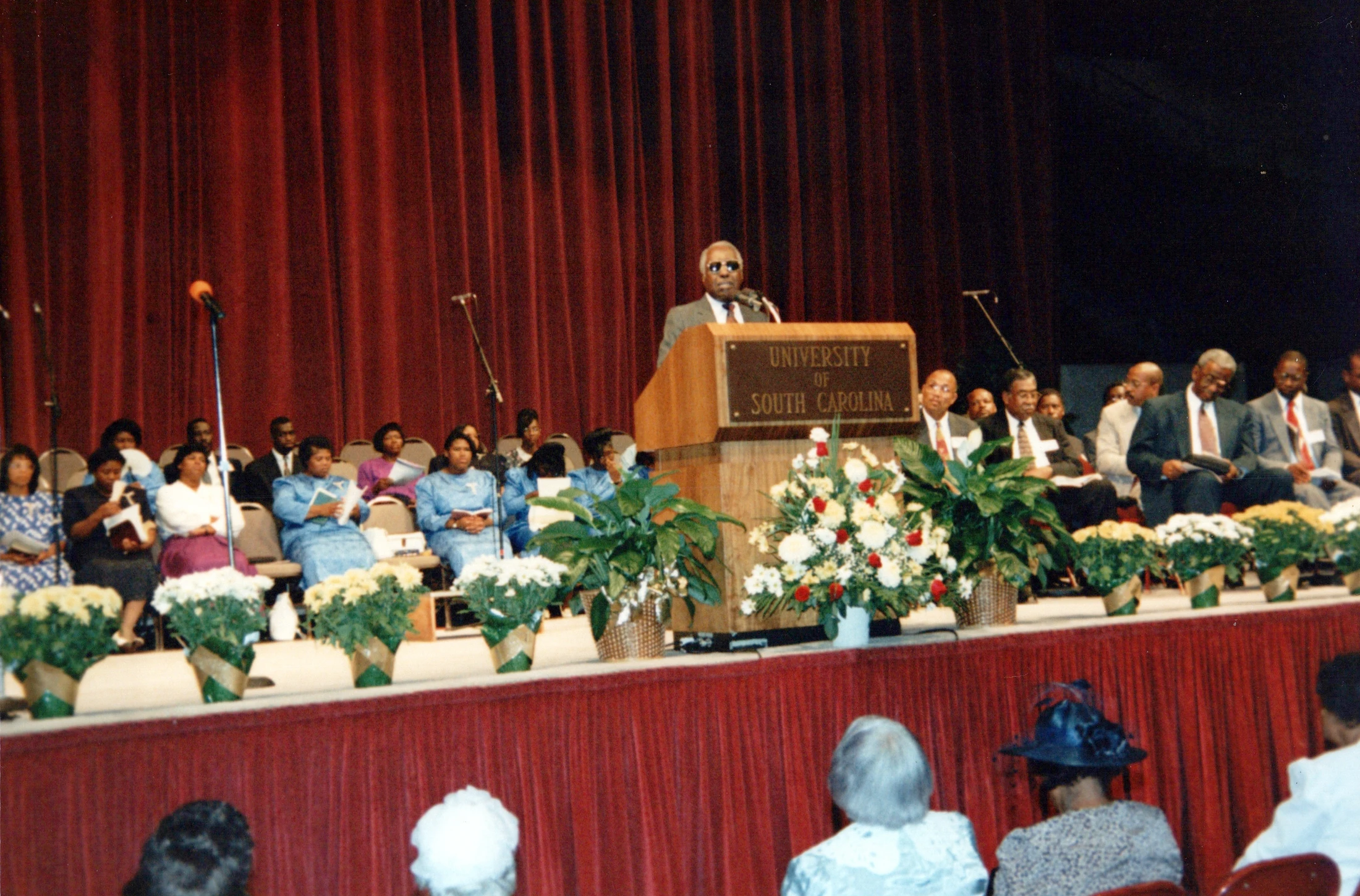 An older Black man speaks at a University of South Carolina podium on a stage. An audience and other participants are seated before a large red curtain.