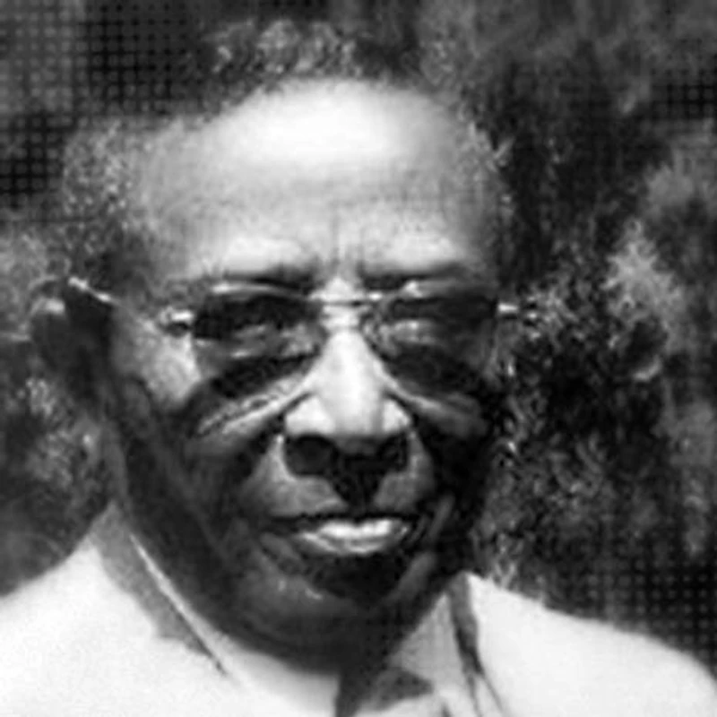 A grainy, black and white close-up portrait of an elderly African American man named Gus Harris. He is wearing glasses and smiling slightly.