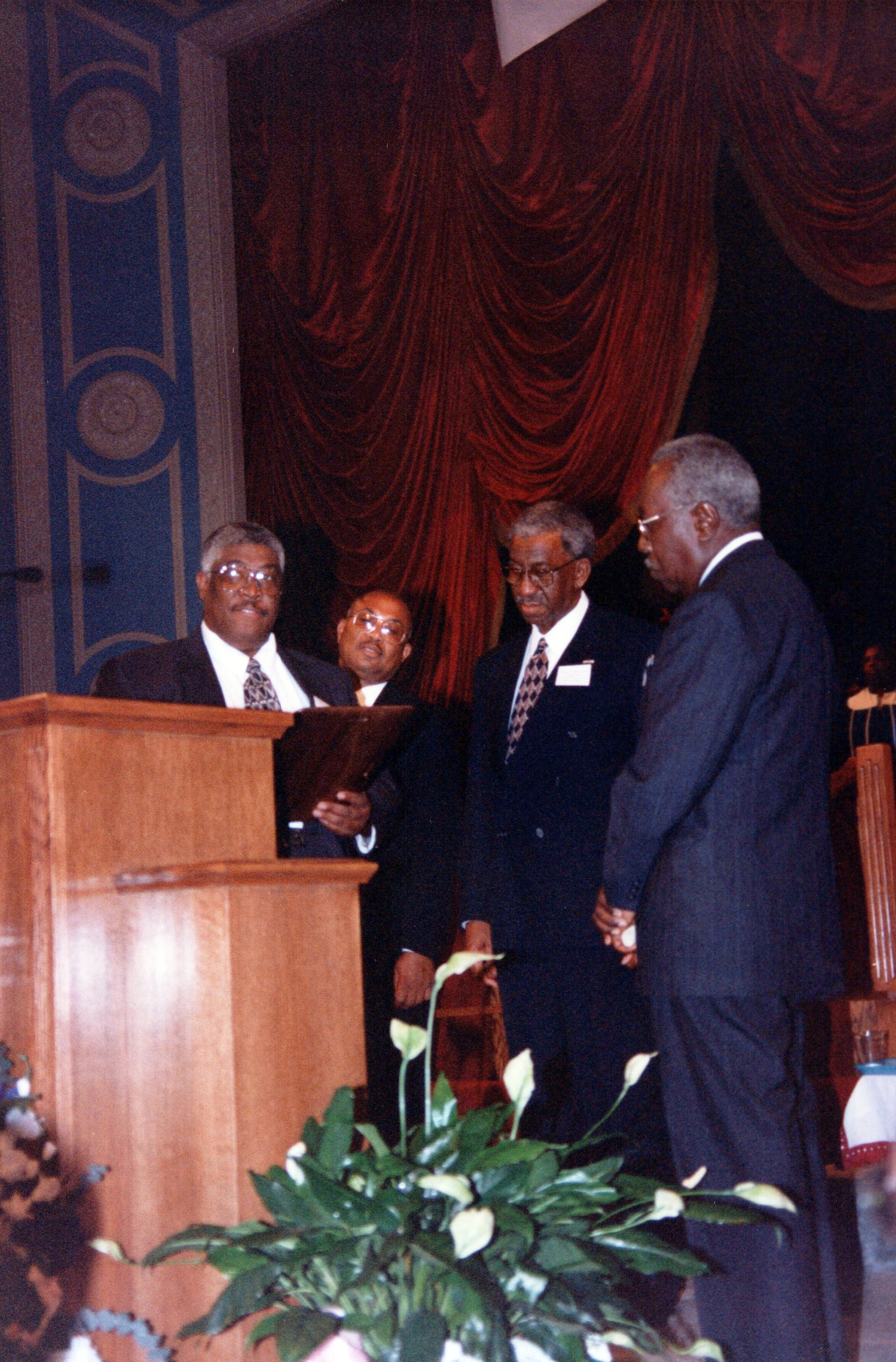 Four older Black men in dark suits stand on a stage. One man is at a wooden podium, speaking, while the others look on. Red curtains are in the background.