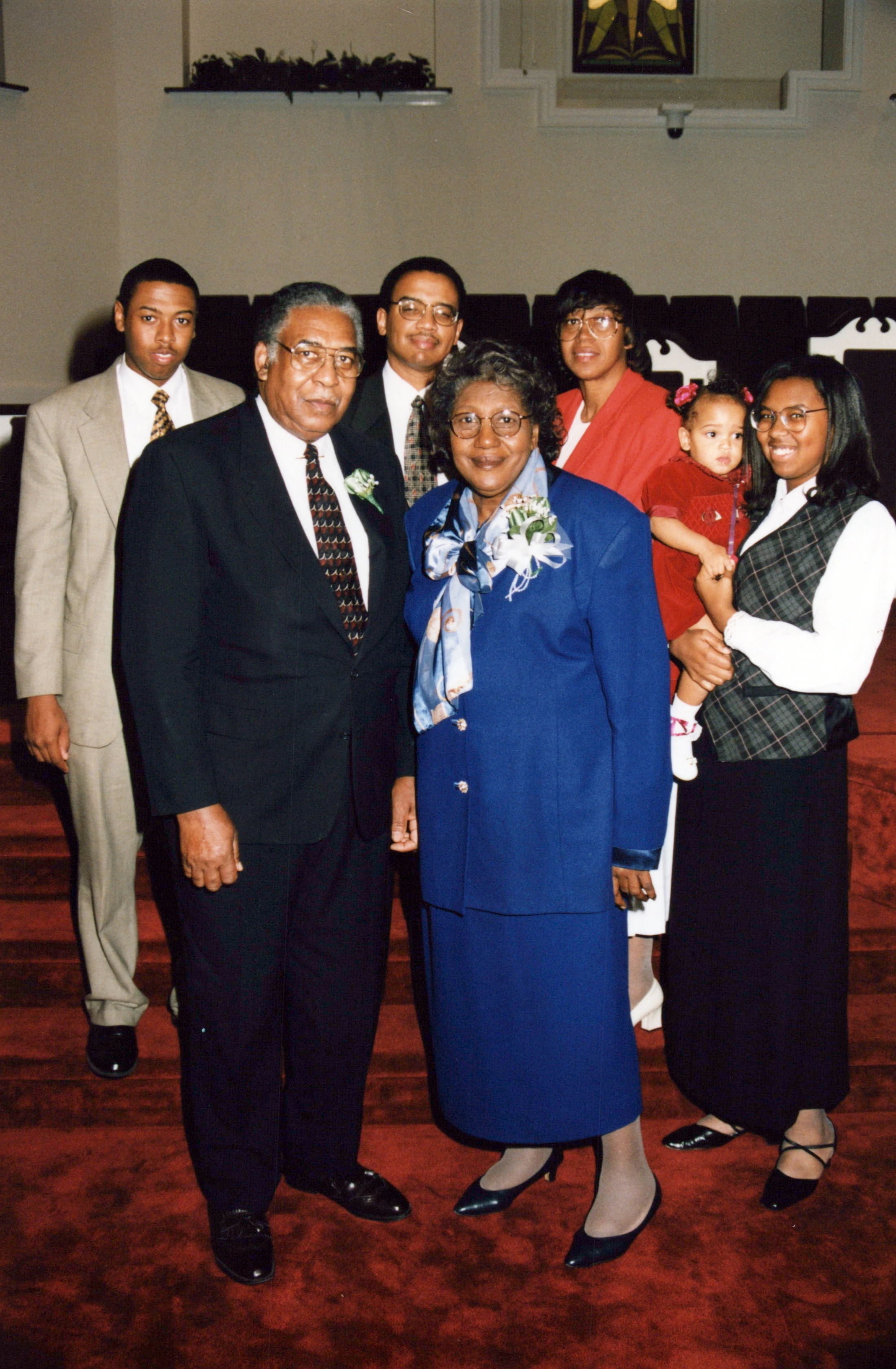 A multi-generational African American family of seven, including a baby, poses for a formal portrait on the red carpeted stairs of a church.