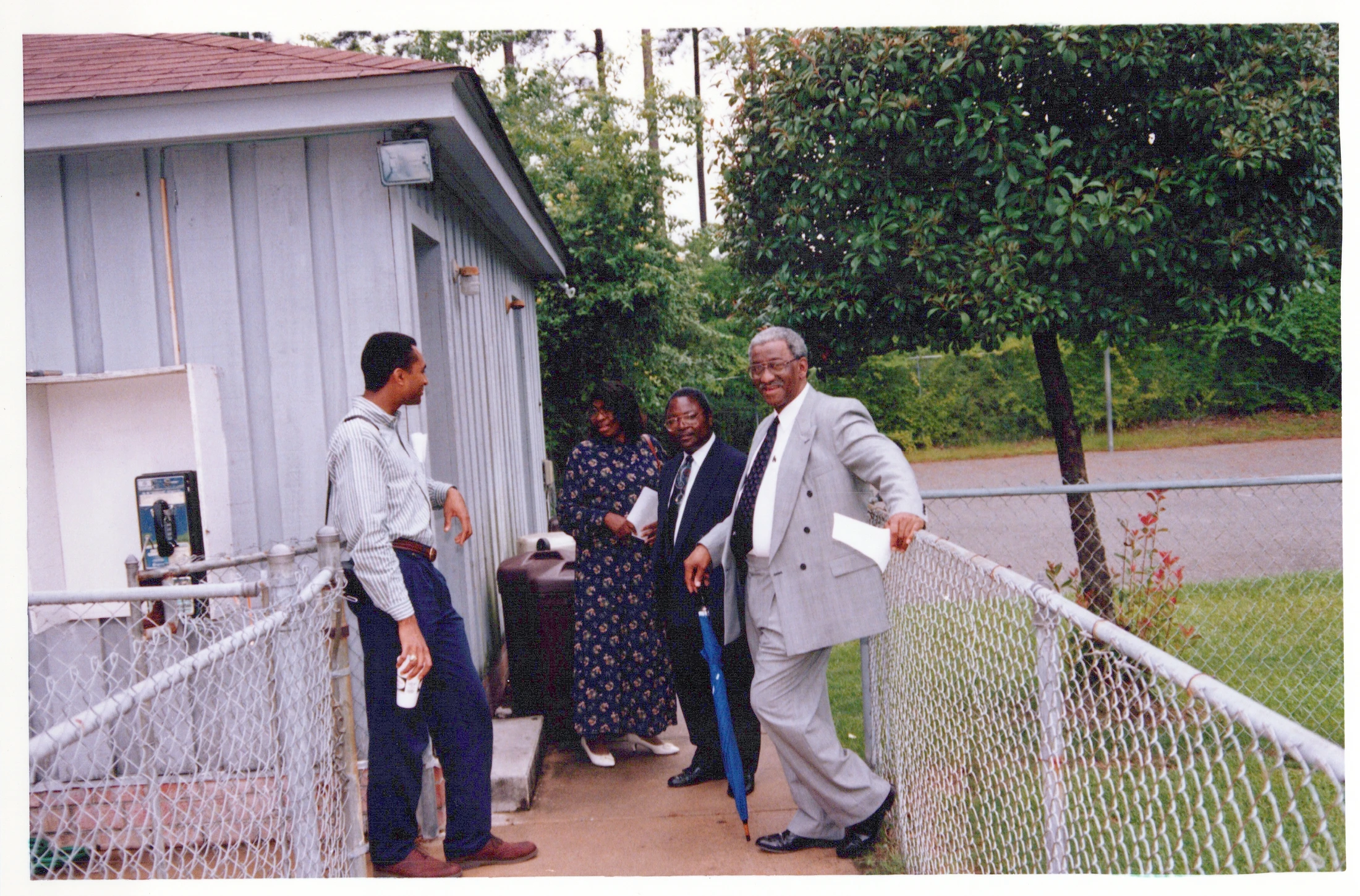 A vintage photo of four Black adults in dress clothes chatting on an outdoor walkway next to a building with a payphone and a chain-link fence.