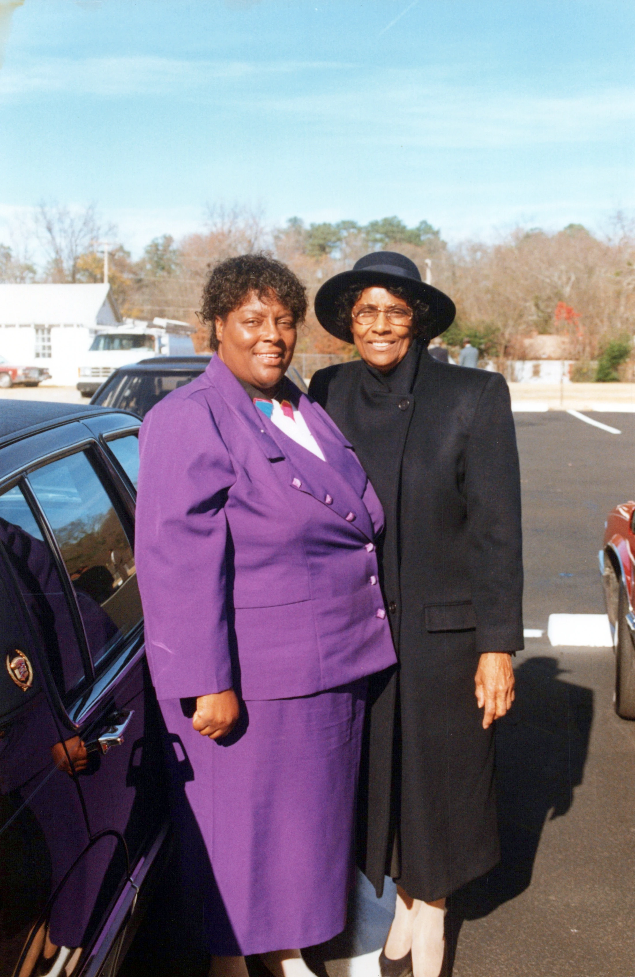 Two smiling Black women pose for a photo in a parking lot. One wears a vibrant purple suit, while the other wears a black coat and hat on a sunny day.