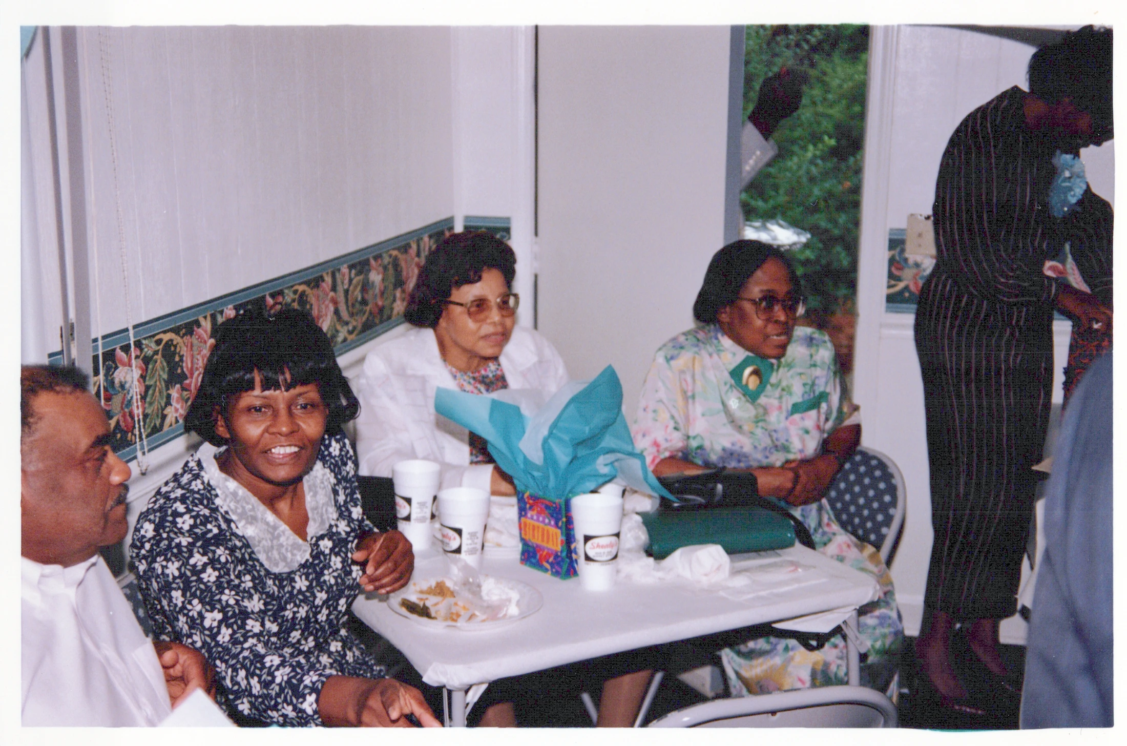 A vintage film photograph of four Black adults sitting around a white table at an indoor gathering, with one woman smiling directly at the camera.