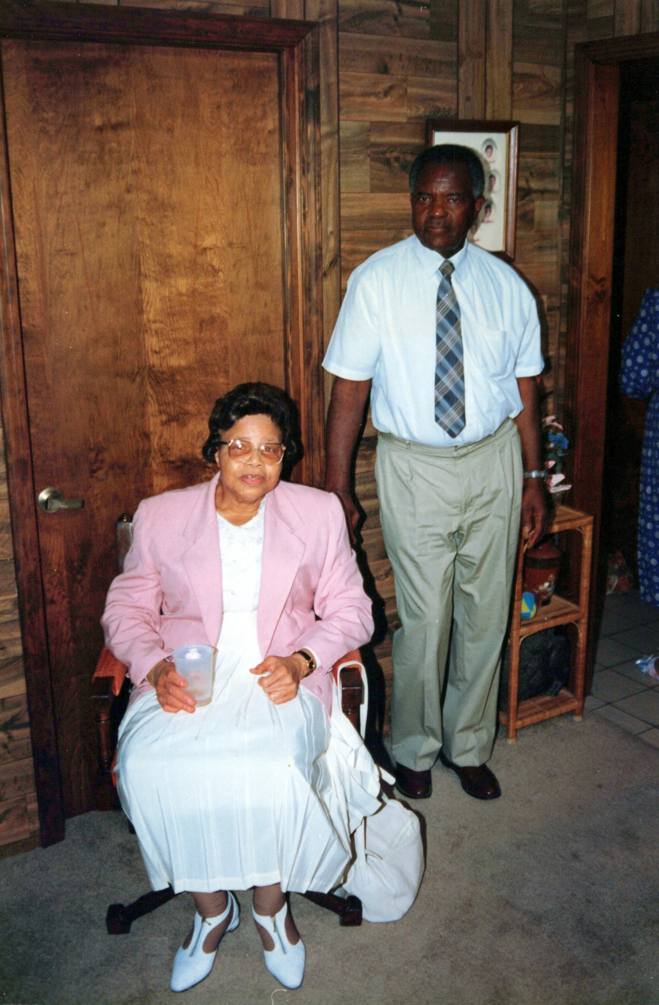 A vintage photograph of an older Black couple. A woman in a pink blazer is seated in a chair while a man in a shirt and tie stands behind her.