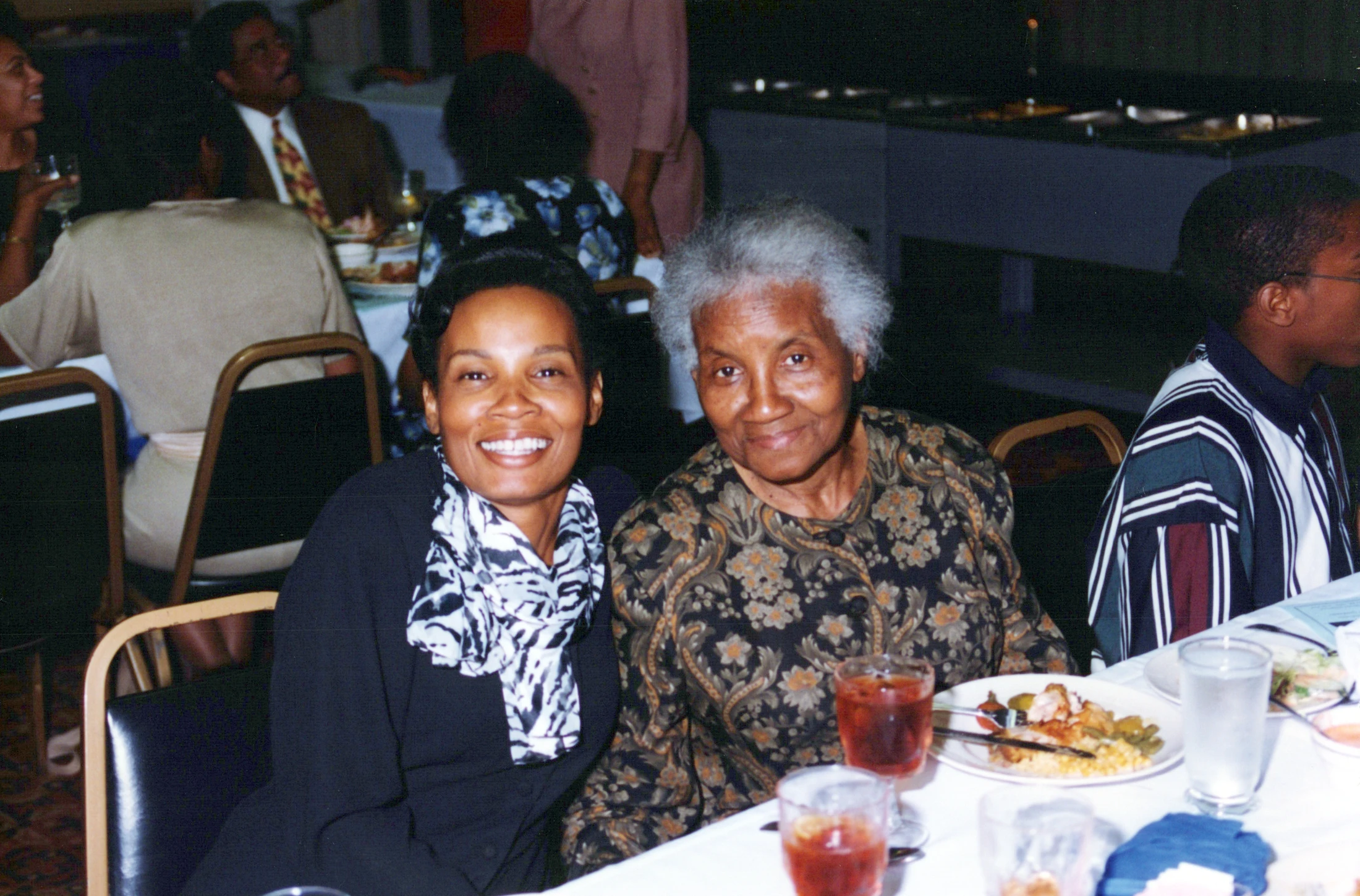 A younger Black woman and an older Black woman sit side-by-side at a dinner table, both smiling warmly for the camera during a gathering.