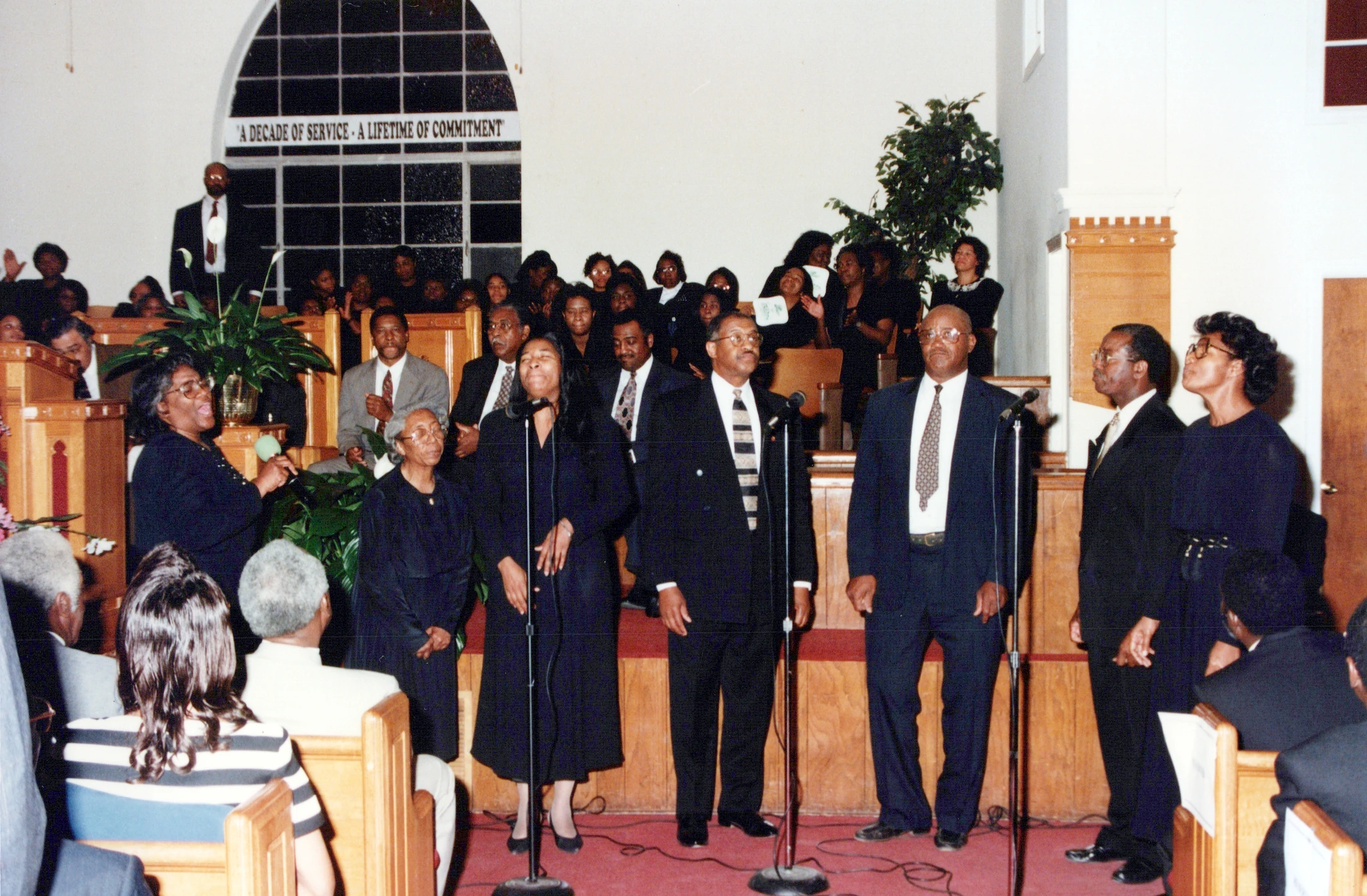 A Black choir of men and women dressed in formal attire sings into microphones at the front of a church during a service for a special anniversary.