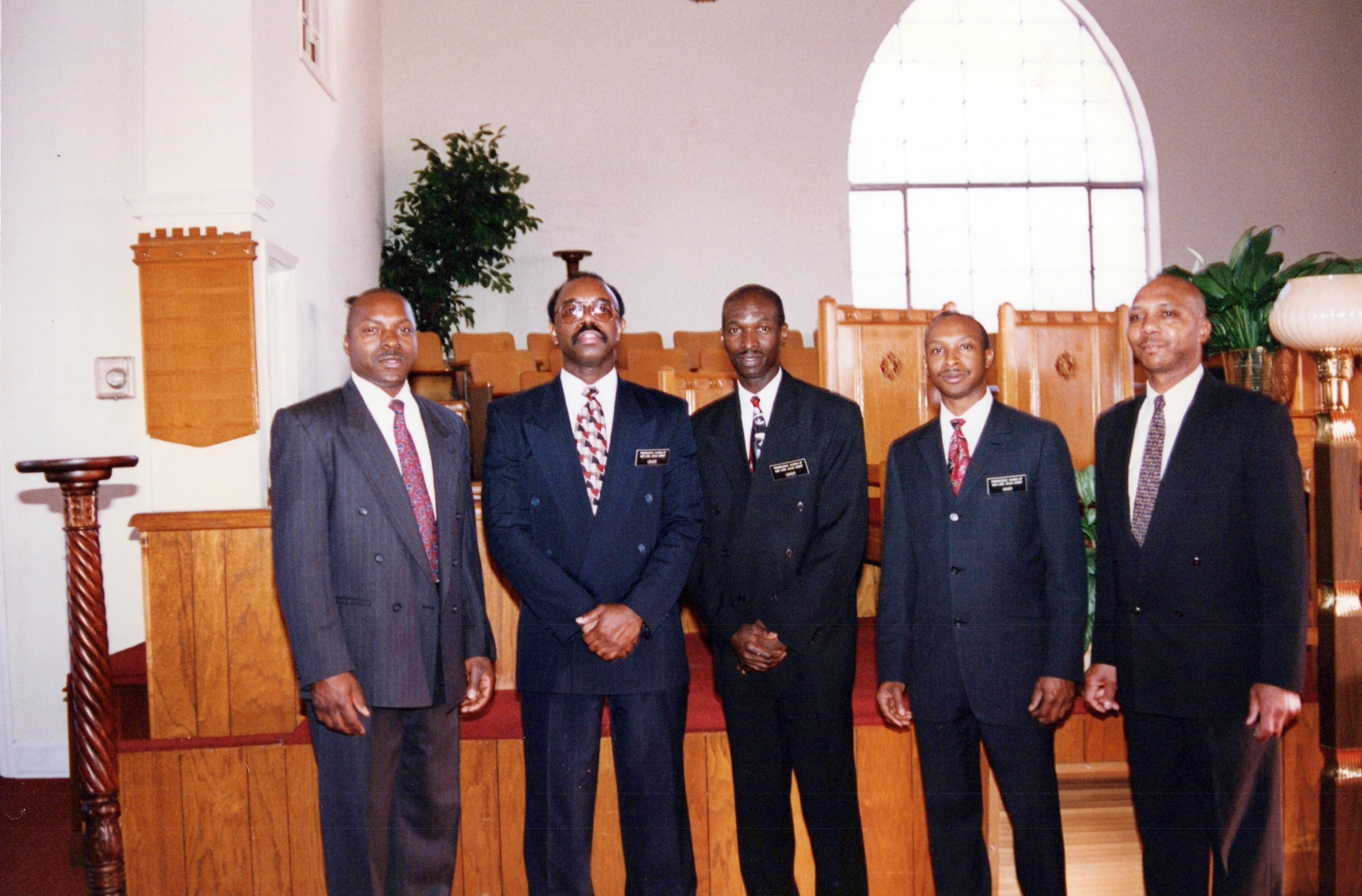 Five African American men in dark suits and ties stand for a group photo inside a church. They are standing in front of wooden pews and an arched window.