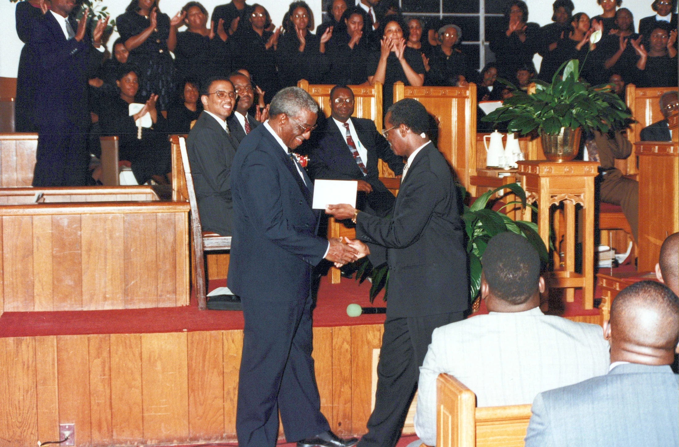 Two Black men in suits shake hands on a stage in a church, as one presents the other with an envelope while the congregation applauds in the background.