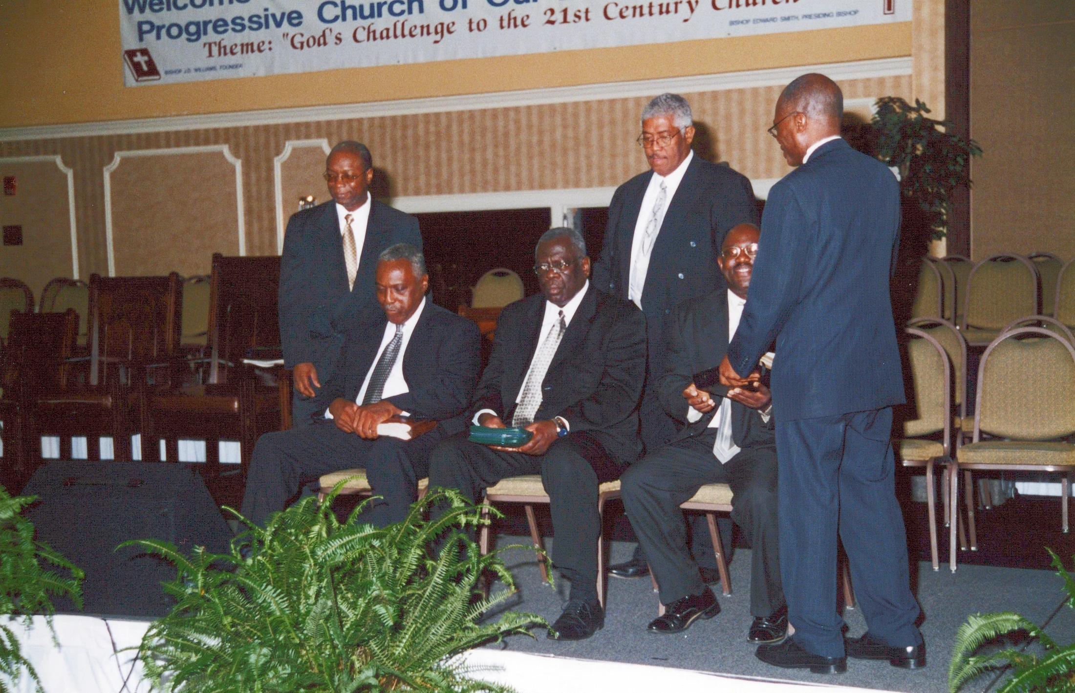 A group of six older African American men in dark suits on a stage at a church event. Some are seated while one man stands, addressing another.