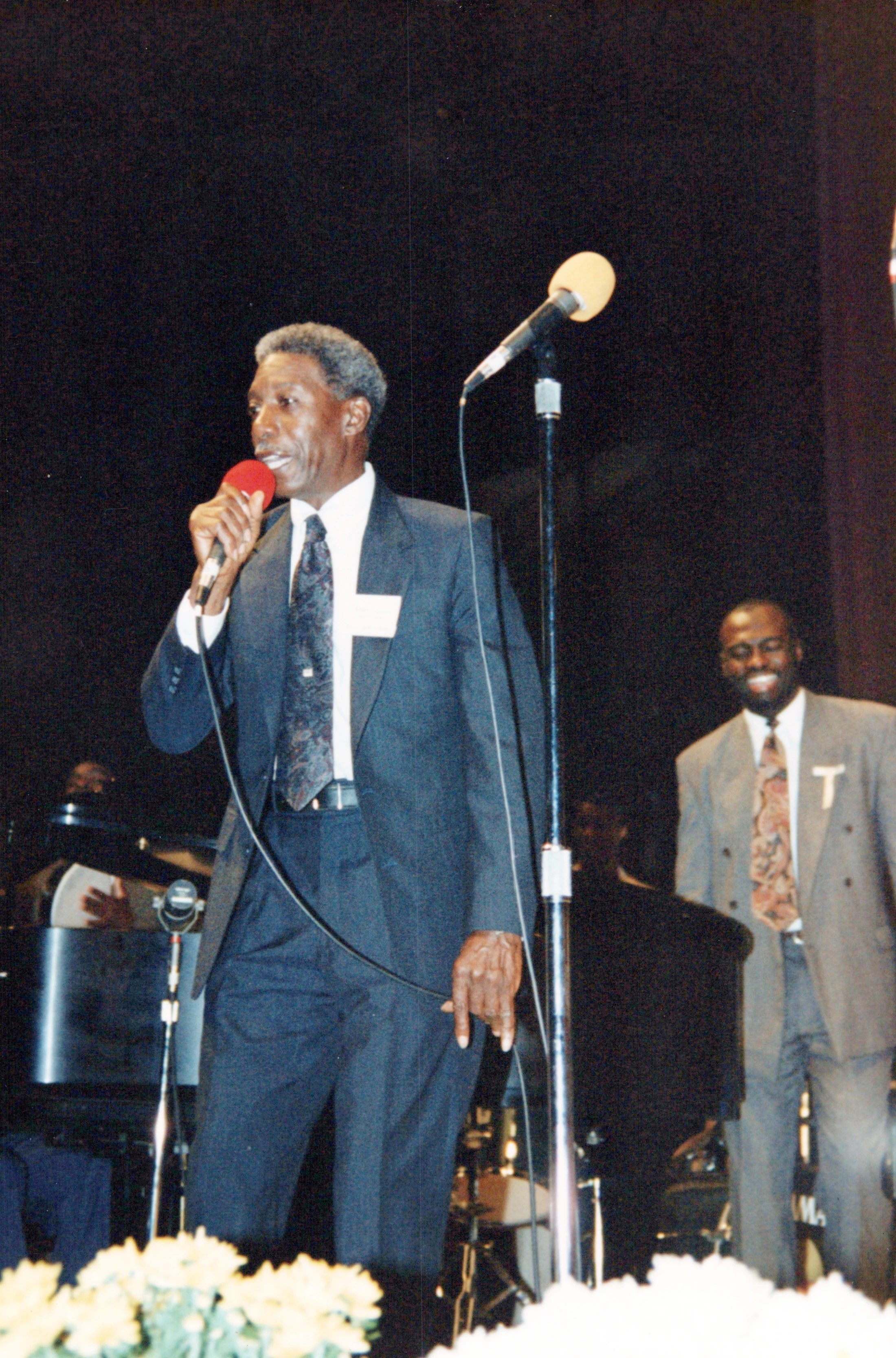 A man with graying hair in a dark suit sings into a red microphone on stage. Another man in a suit smiles in the background, with flowers in the foreground.