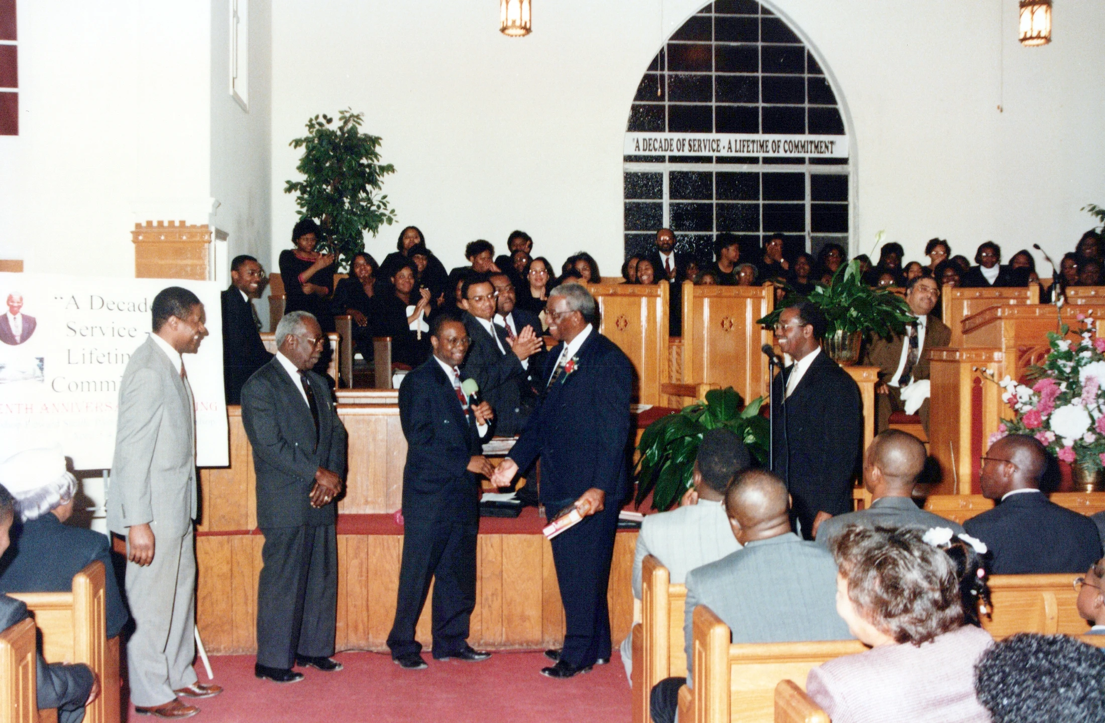 Men in suits stand at the front of a church during a service. One man shakes another's hand as the congregation watches from the wooden pews.