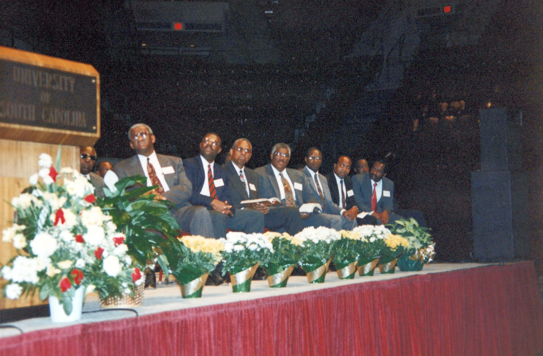 A group of eight African American men in suits and ties are seated in a row on a stage behind a table with floral arrangements.