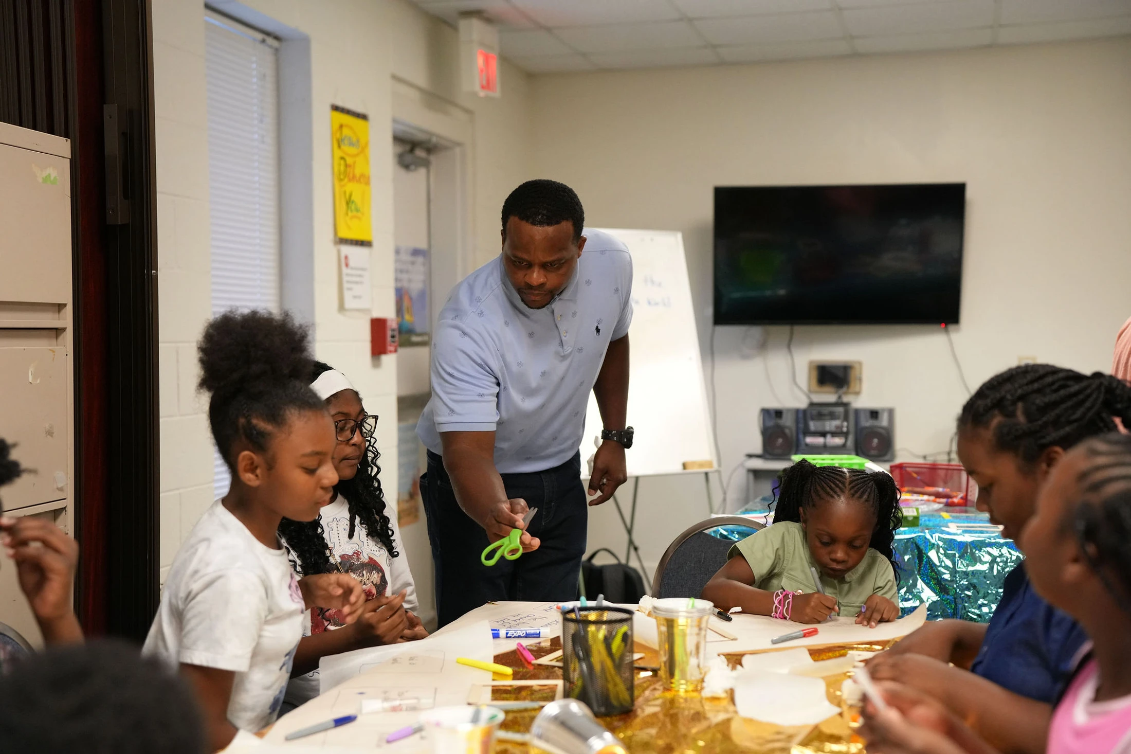 A Black man leans over a table to help a group of young Black girls with an arts and crafts project in a classroom setting.
