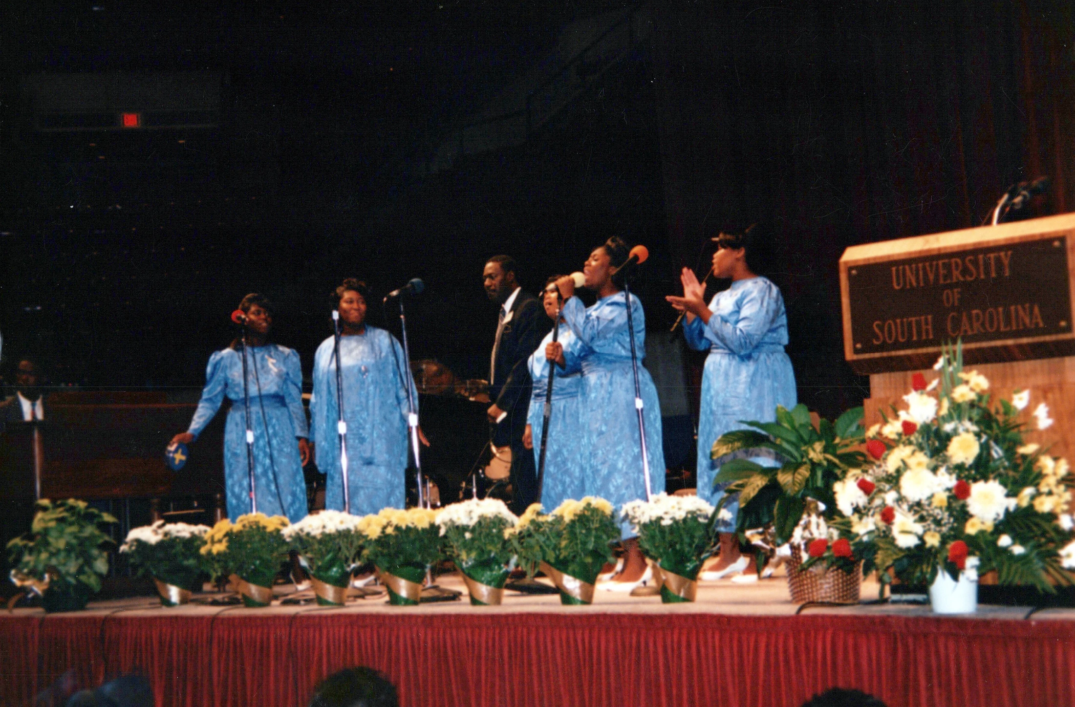 A Black gospel choir of women in matching blue dresses sings on stage at the University of South Carolina, with potted flowers lining the stage.