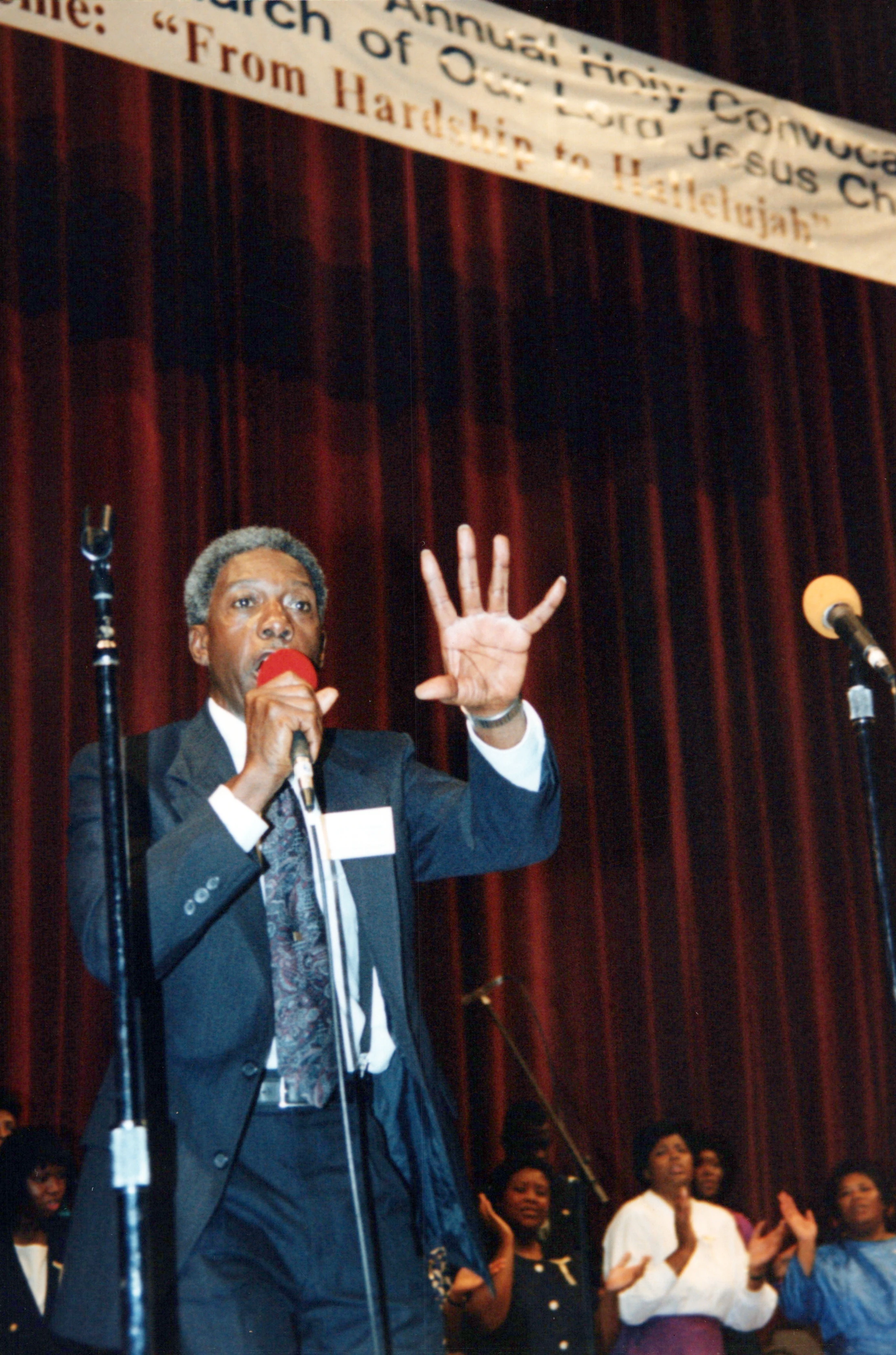 An older Black man in a suit preaches into a microphone on a stage with a red curtain. He gestures with one hand while a choir looks on from behind.