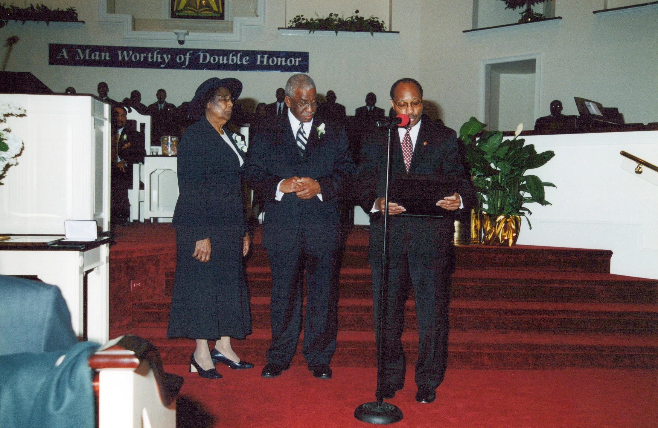 Three formally dressed African American individuals on a red-carpeted stage in a church. A man speaks at a microphone while a man and woman stand beside him.