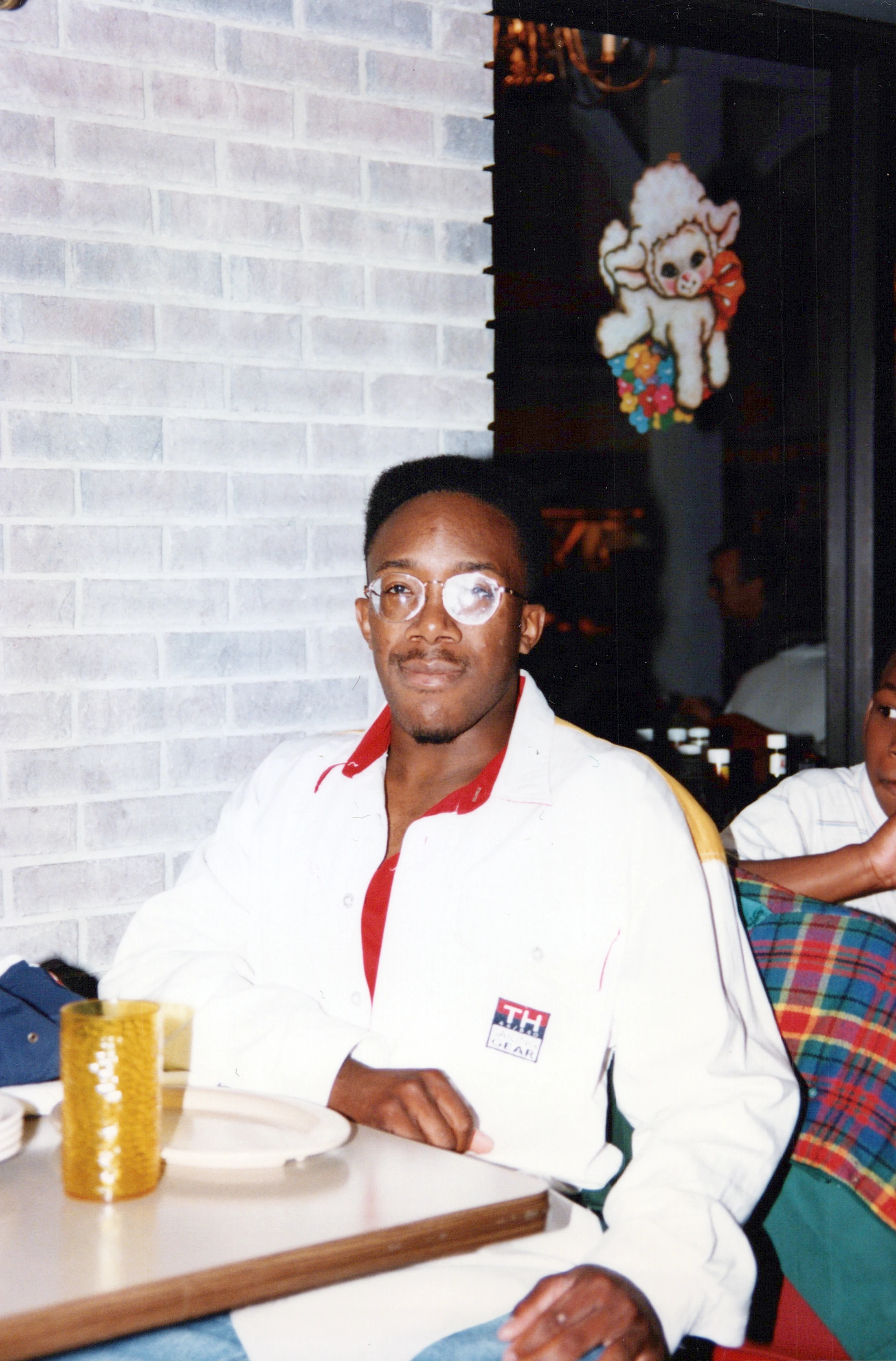A young Black man with a flat top hairstyle and glasses sits at a restaurant table, wearing a white button-up shirt with a red collar over a red shirt.