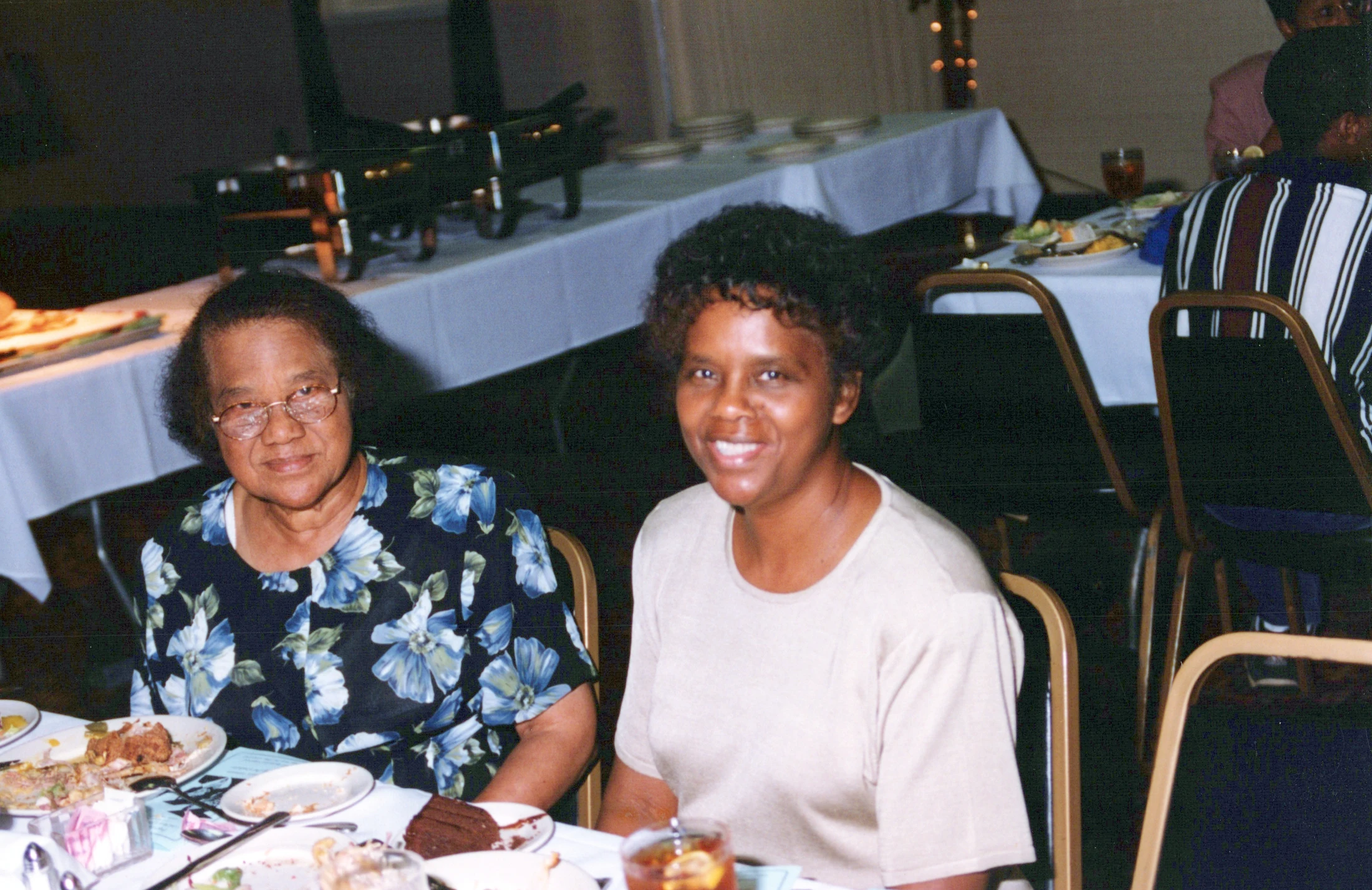 Two African American women sitting side-by-side at a dinner table in a banquet hall. They are both smiling at the camera after finishing their meal.